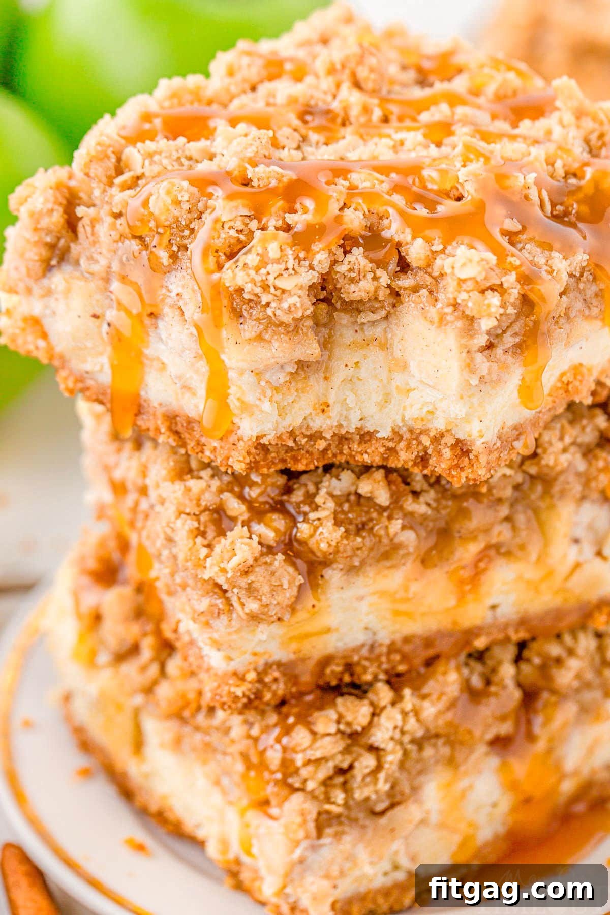 Three homemade Caramel Apple Cheesecake Bars are stacked on a white plate, with a delicious bite taken out of the top bar and golden caramel generously drizzled over the entire stack.