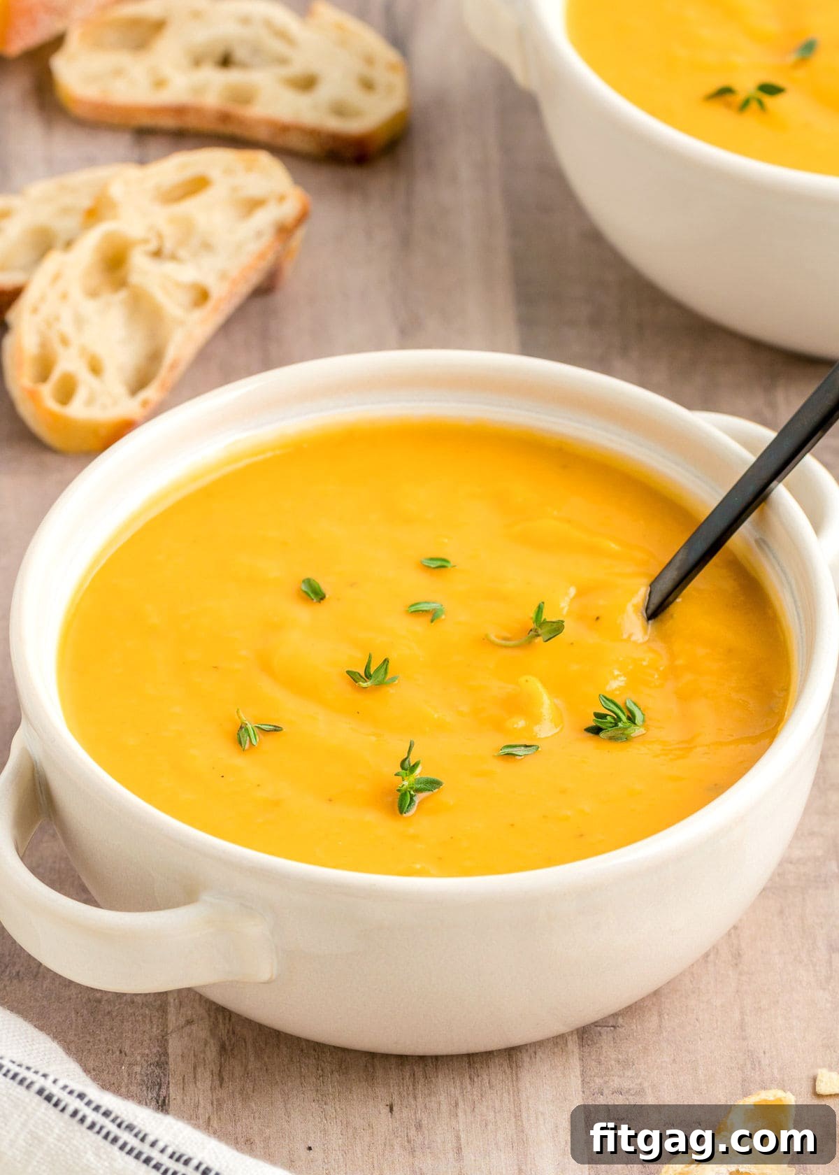 A pristine white round bowl filled with creamy butternut squash soup, elegantly garnished with fresh thyme sprigs. A black spoon rests invitingly in the bowl, ready to be enjoyed.