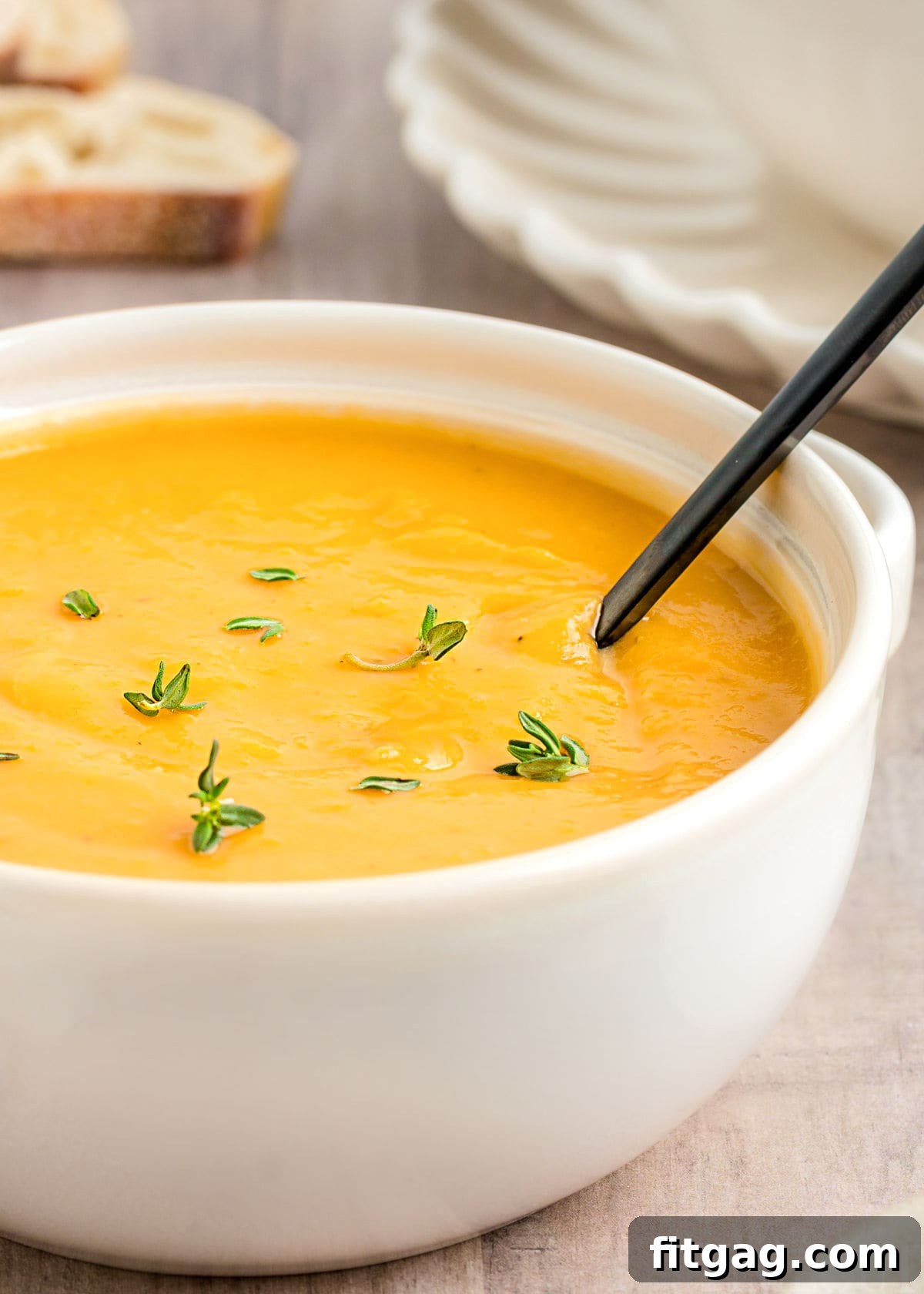 A close-up view of vibrant, creamy butternut squash soup with its bright orange color, served in a beautiful white bowl and garnished with fresh thyme sprigs.