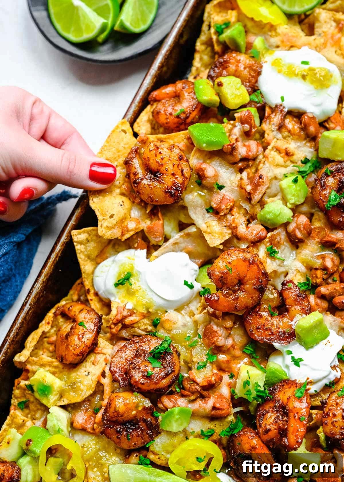 Fiesta Shrimp Crunch 2 A vibrant platter of shrimp nachos on a baking sheet, generously topped with fresh ingredients like avocado, sour cream, and cilantro. A hand reaches in to grab a loaded chip, showcasing the melted cheese and succulent shrimp.