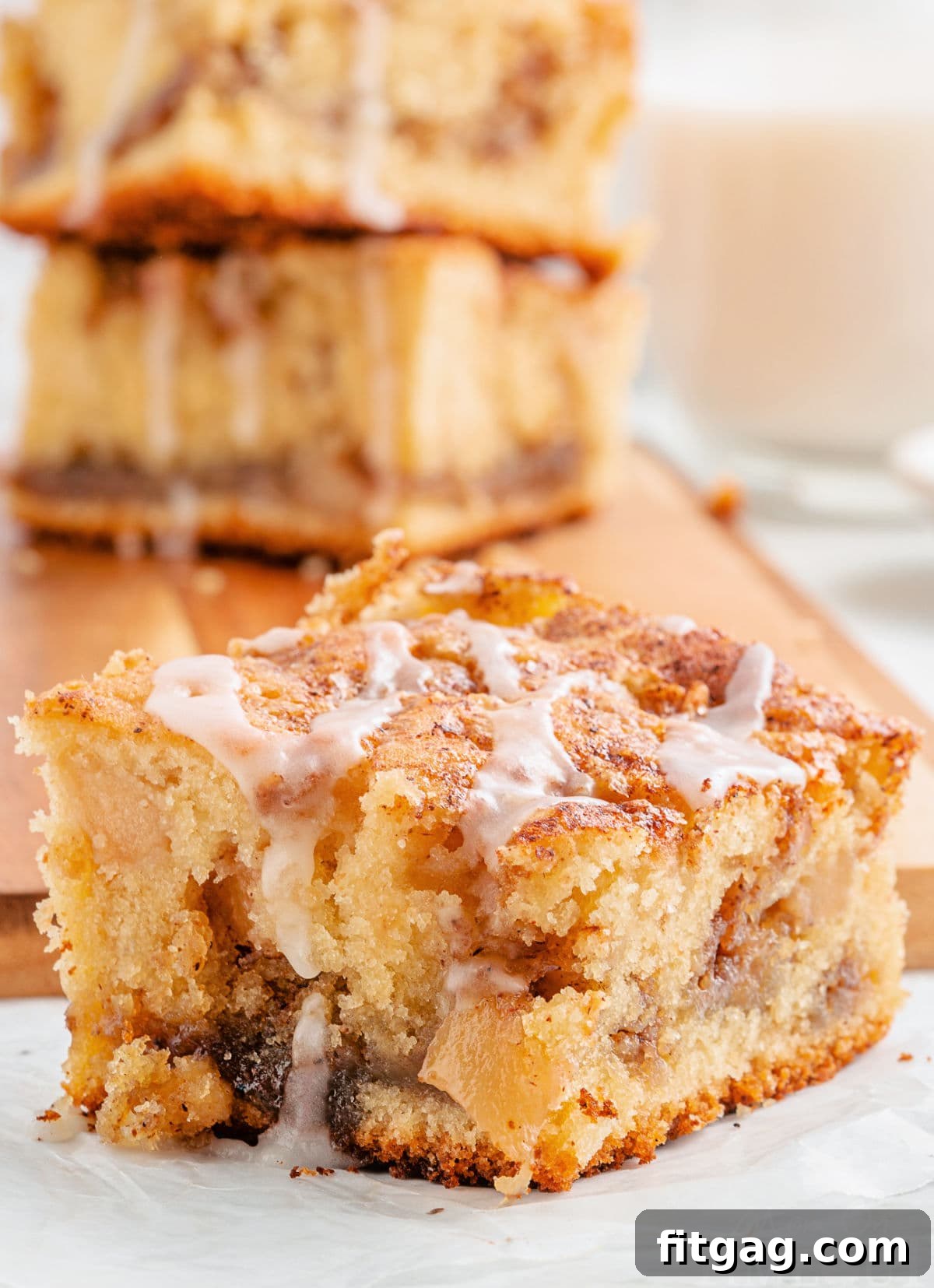 Autumn Apple Coffee Delight 2 one piece of apple coffee cake sitting in front of a cutting board with three more pieces stacked together. coffee cake has a little glaze drizzled over the top.