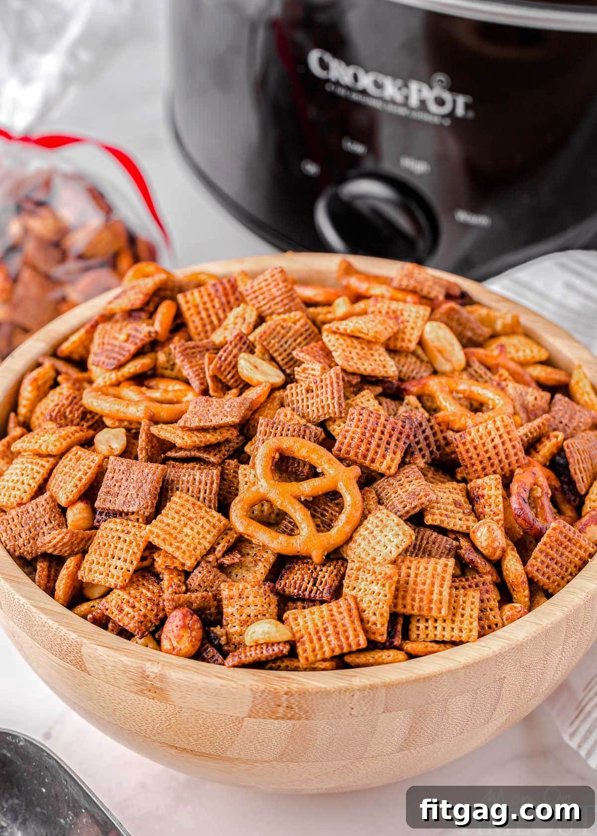 Slow Cooker Chex Mix Perfection 6 A rustic wooden bowl overflowing with crunchy Crockpot Chex Mix placed in front of a sleek black slow cooker, emphasizing the convenience of the slow-cooking method.