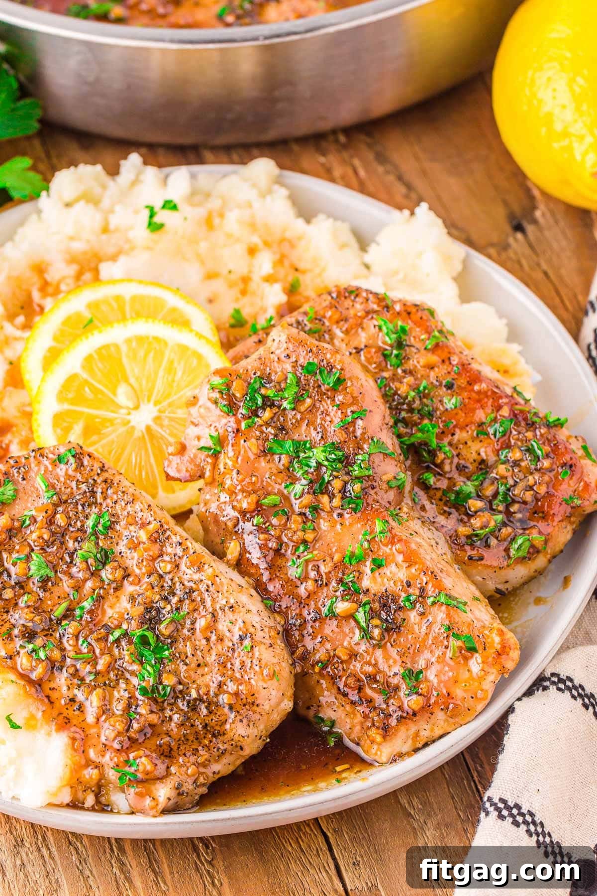 Three perfectly seared honey garlic pork chops resting on a bed of creamy mashed potatoes, garnished with fresh parsley and vibrant lemon slices. The plate is beautifully presented on a rustic wooden board, highlighting the dish's irresistible appeal.