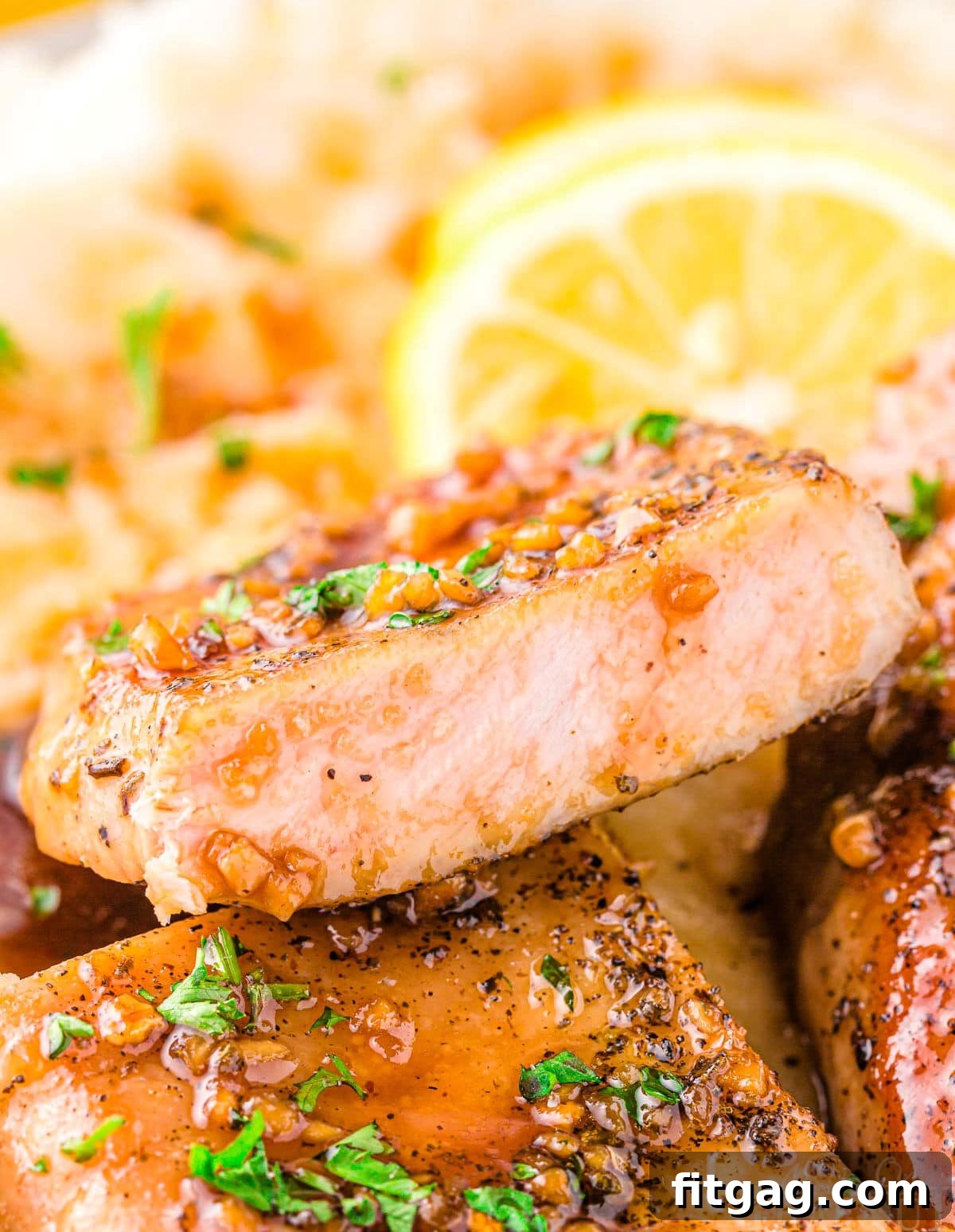 A perfectly cooked honey garlic pork chop, sliced in half, revealing its tender interior and glistening with a rich honey garlic glaze. Fresh parsley garnishes the chop, with vibrant sliced lemons visible in the soft background, emphasizing the dish's fresh and appetizing presentation.