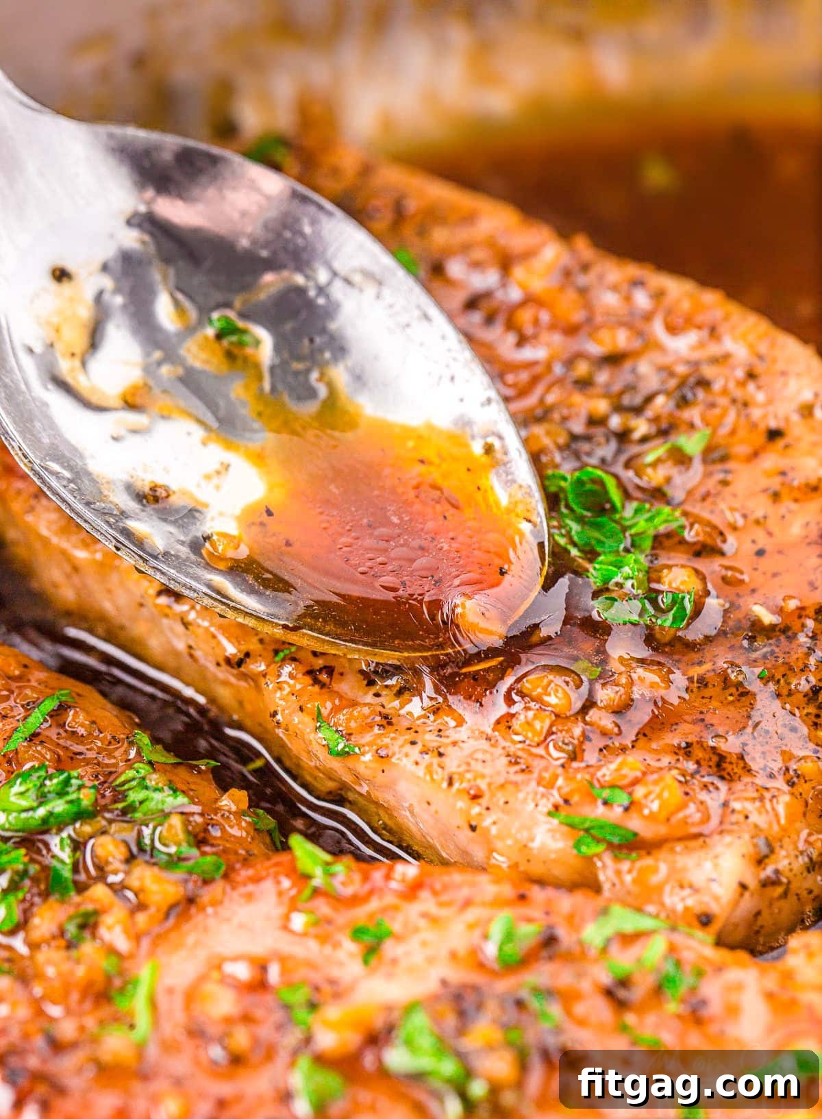 A close-up shot of a spoonful of glistening honey garlic glaze being expertly spooned onto a seared pork chop in a hot skillet, showcasing the rich texture and appealing shine of the sauce.