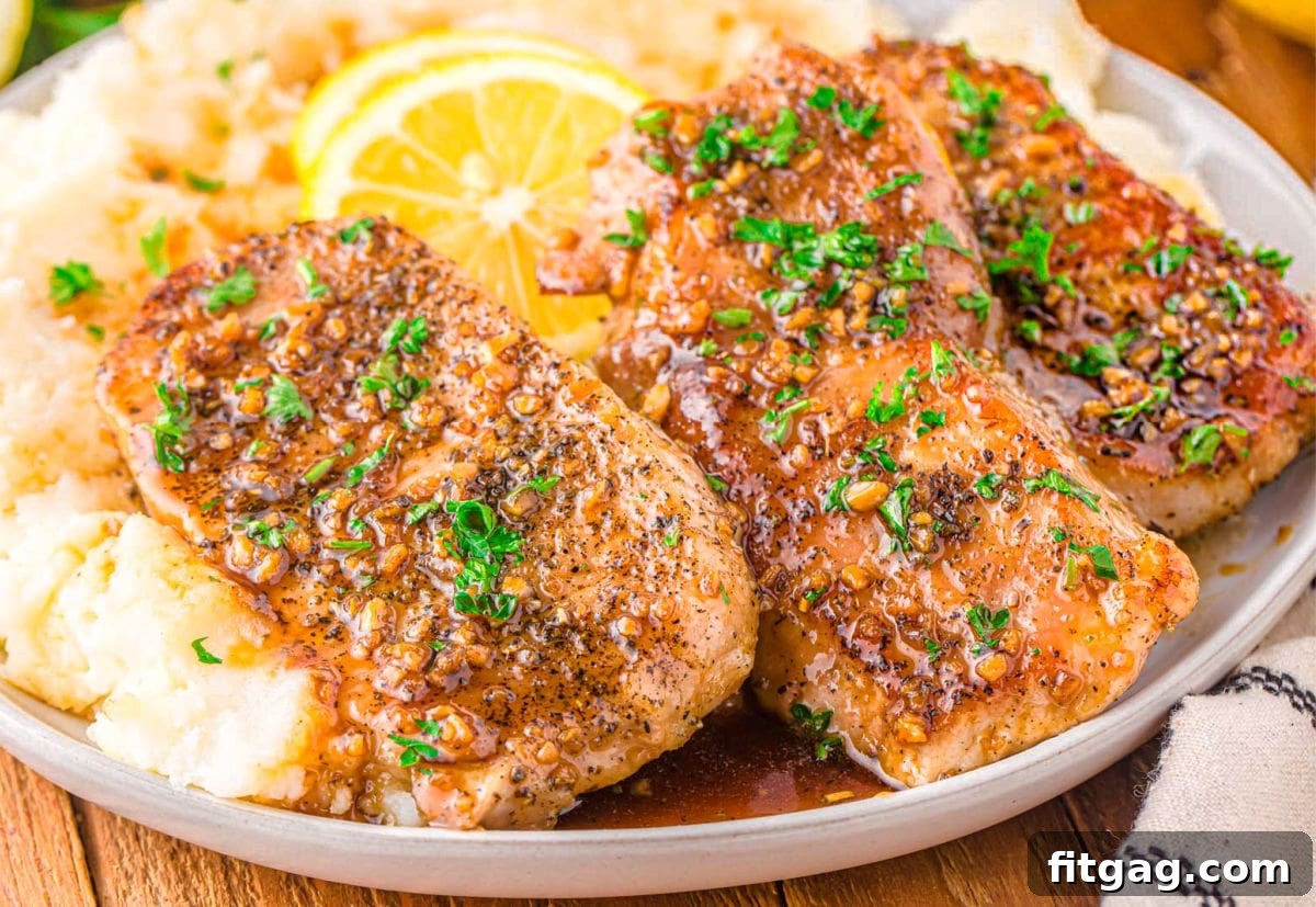 Three perfectly cooked honey garlic pork chops, glistening with sauce, artfully arranged on a bed of fluffy mashed potatoes. The dish is elegantly garnished with fresh parsley and bright lemon slices, presented on a rustic wooden board, ready to be enjoyed.
