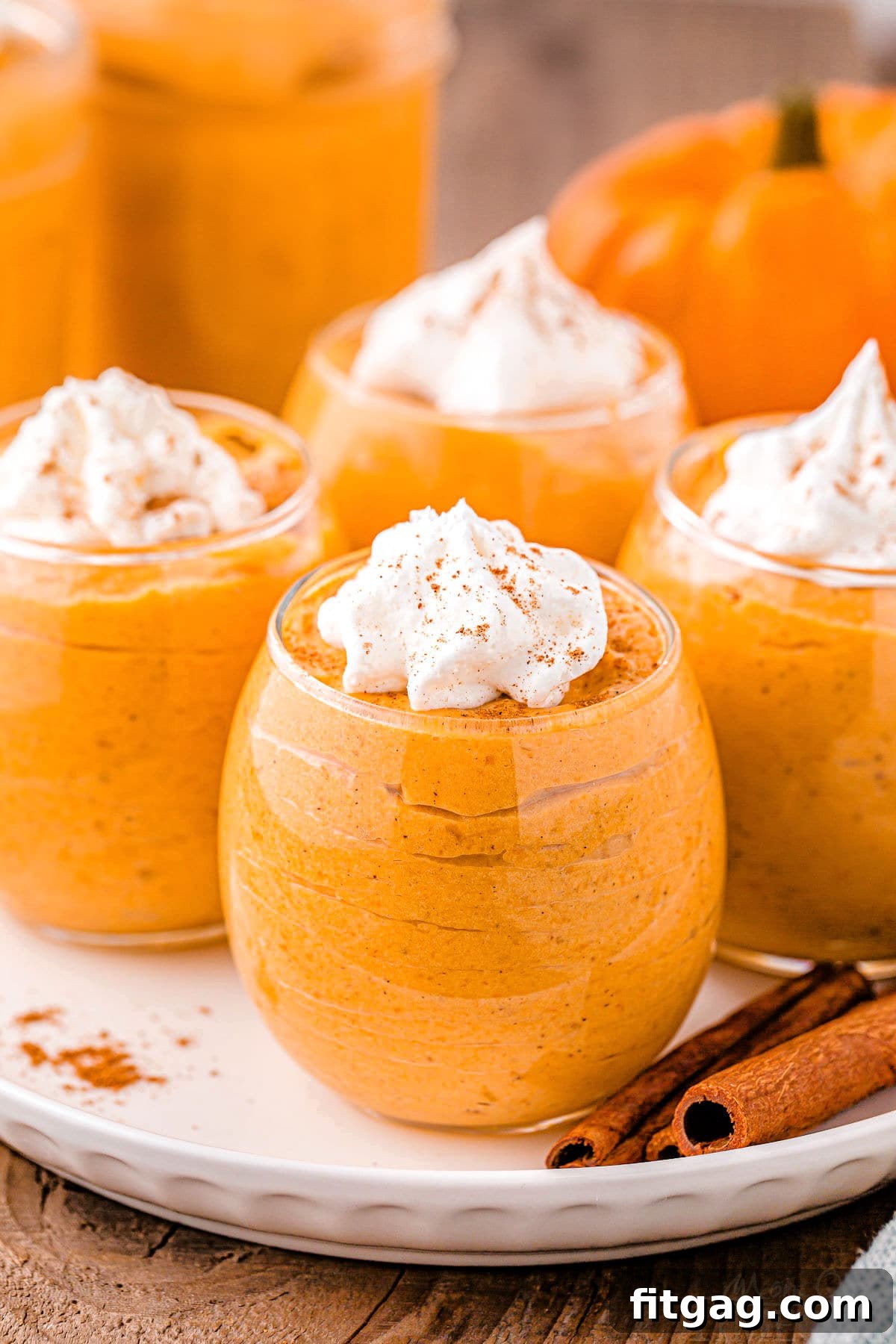 Creamy Pumpkin Cloud 2 four short wine glasses filled with pumpkin mousse, topped with whipped cream and pumpkin pie spice. the glasses are sitting on a round white plate on a wood surface. there are two cinnamon sticks on the plate.