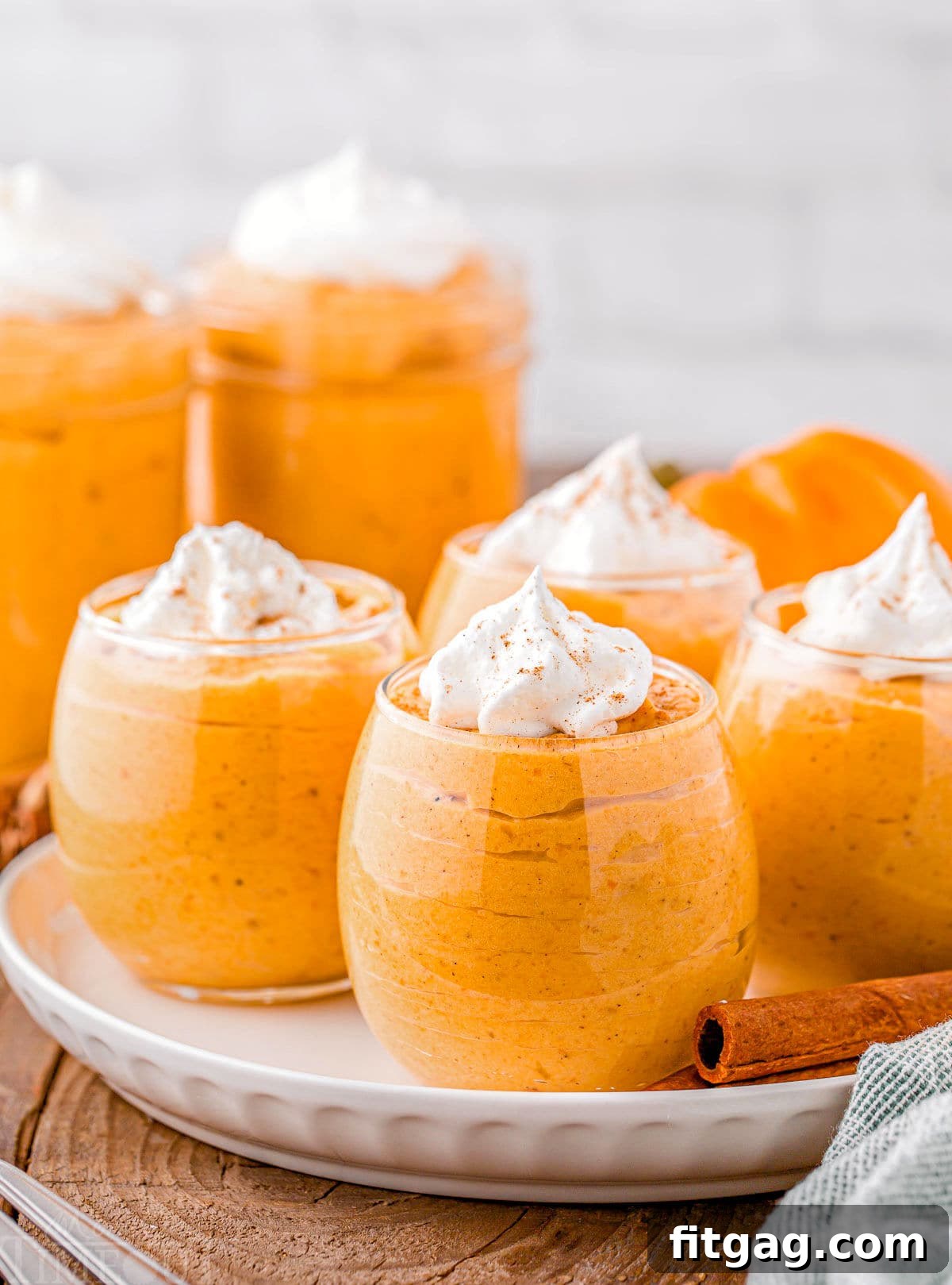Creamy Pumpkin Cloud 6 four short wine glasses filled with pumpkin mousse, topped with whipped cream and pumpkin pie spice. the glasses are sitting on a round white plate on a wood surface. there are two cinnamon sticks on the plate.
