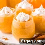 Creamy Pumpkin Cloud 7 four stemless wine glasses filled with pumpkin mousse, topped with whipped cream and pumpkin pie spice. the glasses are sitting on a round white plate on a wood surface. there are two cinnamon sticks on the plate.