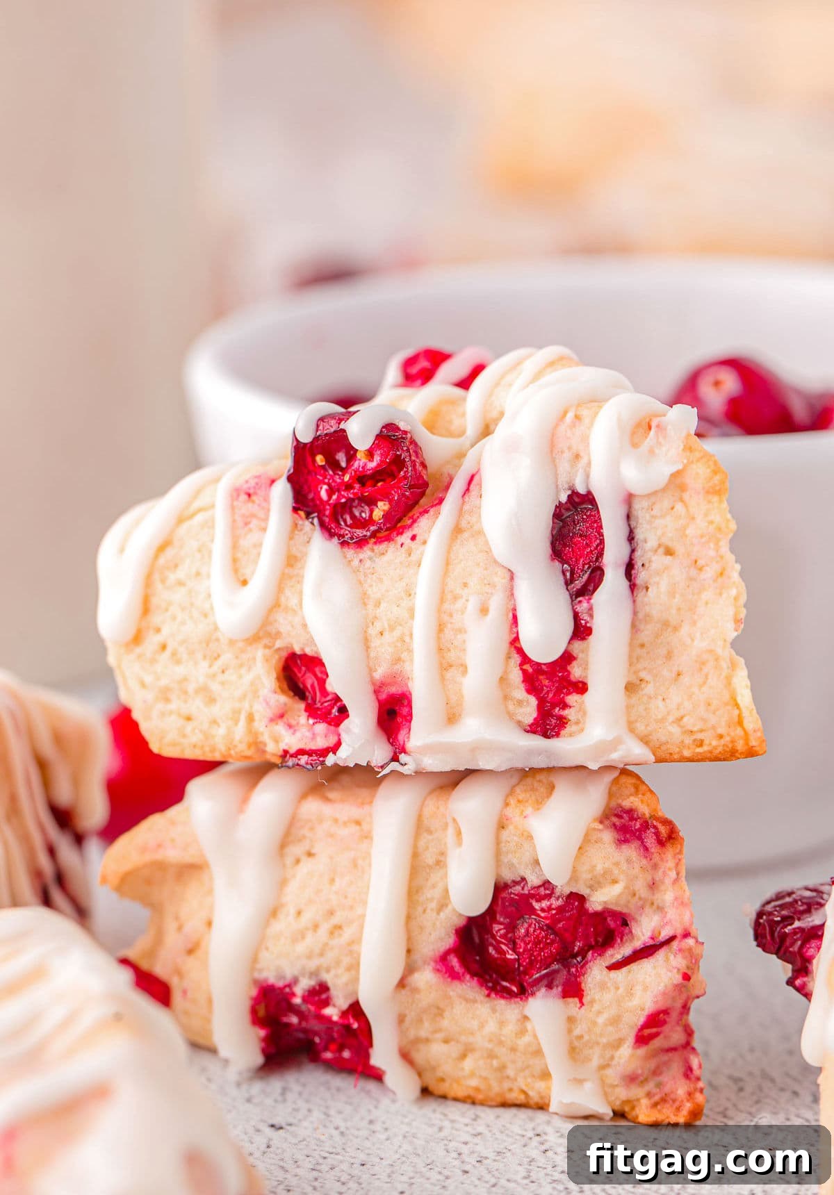 Tangy Cranberry Scones 2 two small cranberry scones stacked with a simple glaze drizzled on top. a small white bowl of cranberries are in back as well as more scones to the sides.