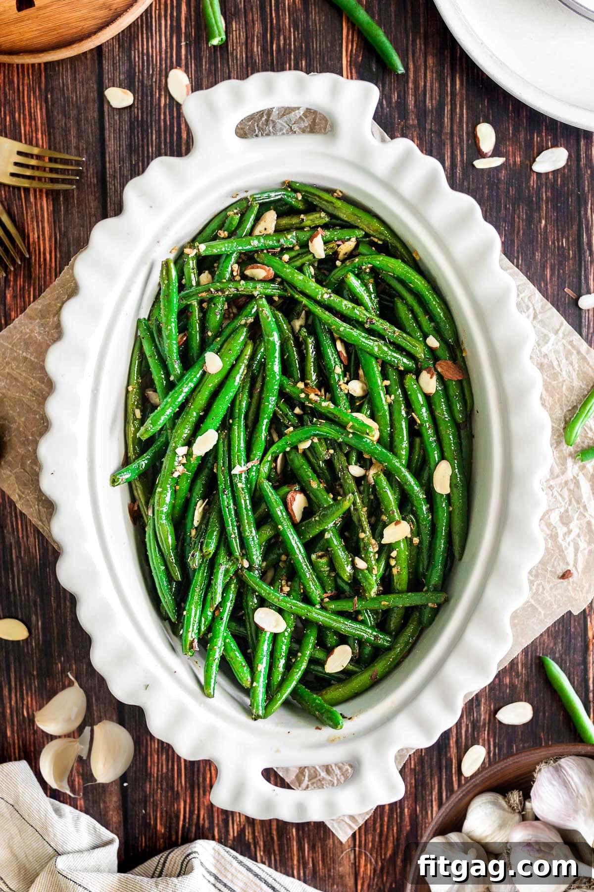 Top-down view of a large white oval serving bowl filled with bright green garlic green beans, elegantly arranged on a dark wood surface.
