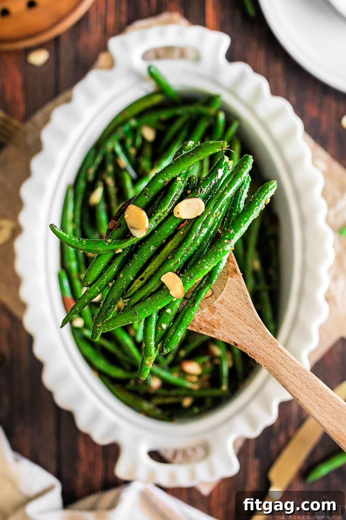 Wooden spoon lifting out a serving of garlic green beans from an oval white serving dish.
