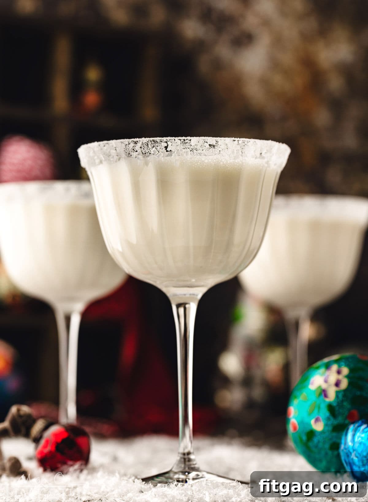Three white chocolate martinis in tall glasses, elegantly dusted with powdered sugar to create a snowy effect. The cocktails are artfully arranged on a pristine snowy surface, surrounded by delicate glass ornaments, evoking a serene winter wonderland.