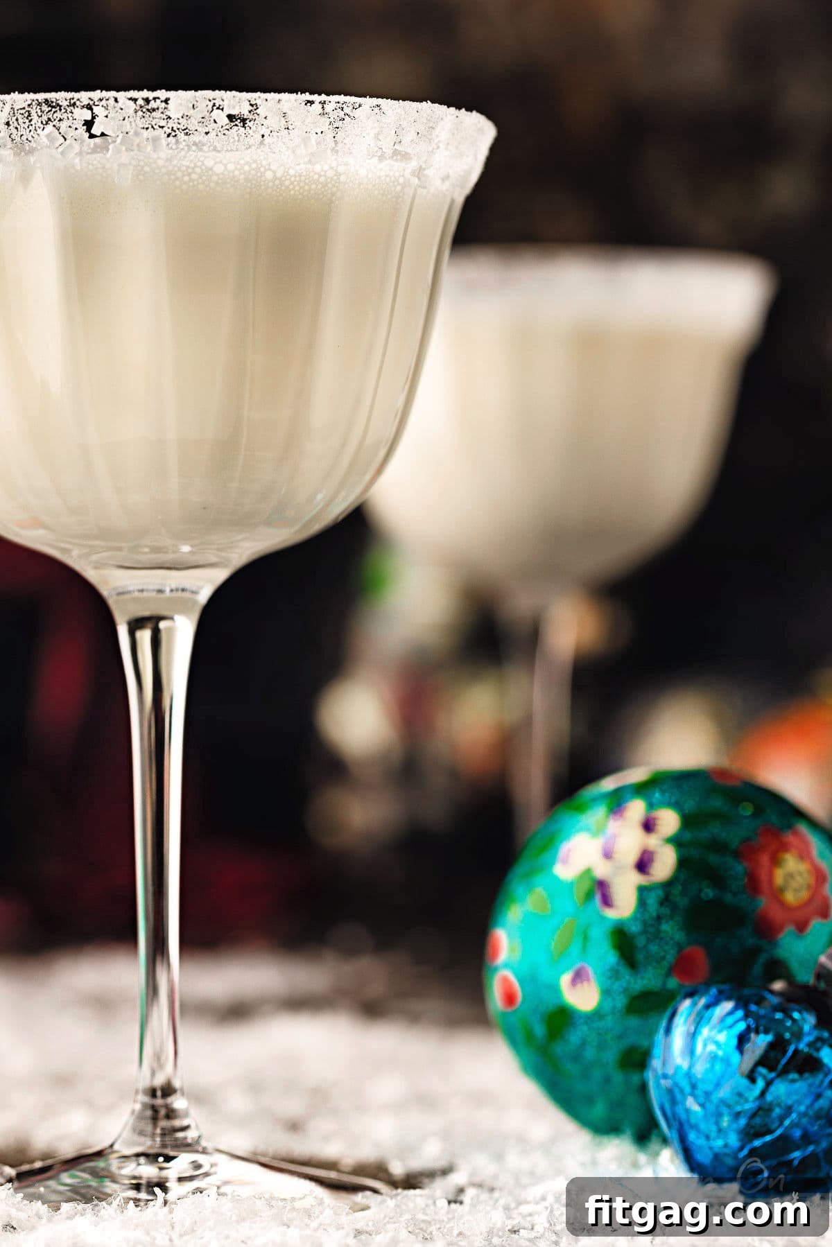 Two white martinis in tall glasses, dusted with powdered sugar for a picturesque snowy aesthetic. The cocktails are placed beside vintage ornaments, with bottles of premium vodka and white chocolate liqueur subtly visible in the soft background, hinting at the key ingredients.