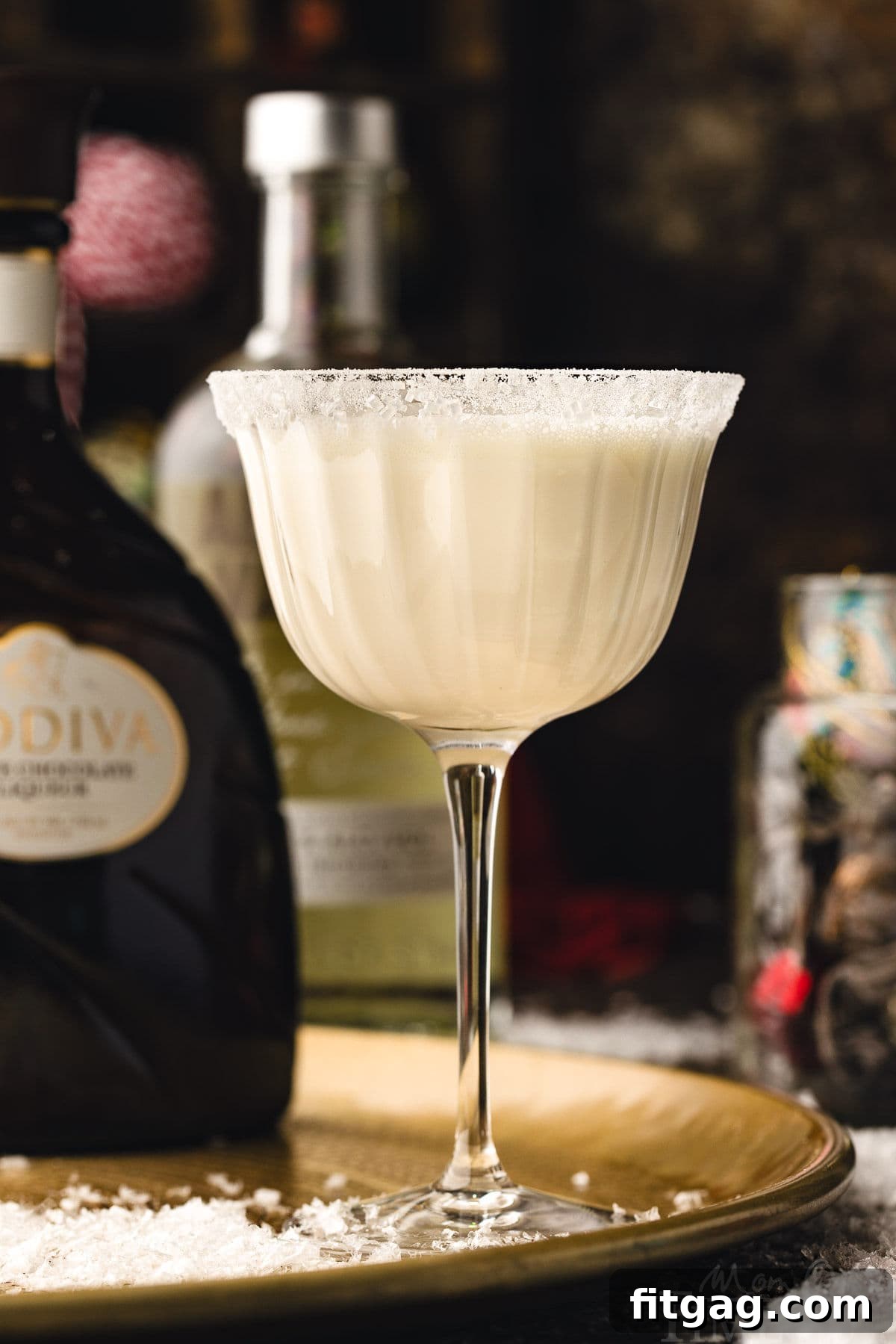 white chocolate martini cocktail in tall glass dusted with powdered sugar for a snowy look. the cocktails is sitting on a gold tray. bottles of vodka and white chocolate liqueur can be seen in the background.