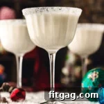 Three white chocolate martinis in tall glasses dusted with powdered sugar for a snowy look. The cocktails are sitting on a snowy surface with glass ornaments scattered about.