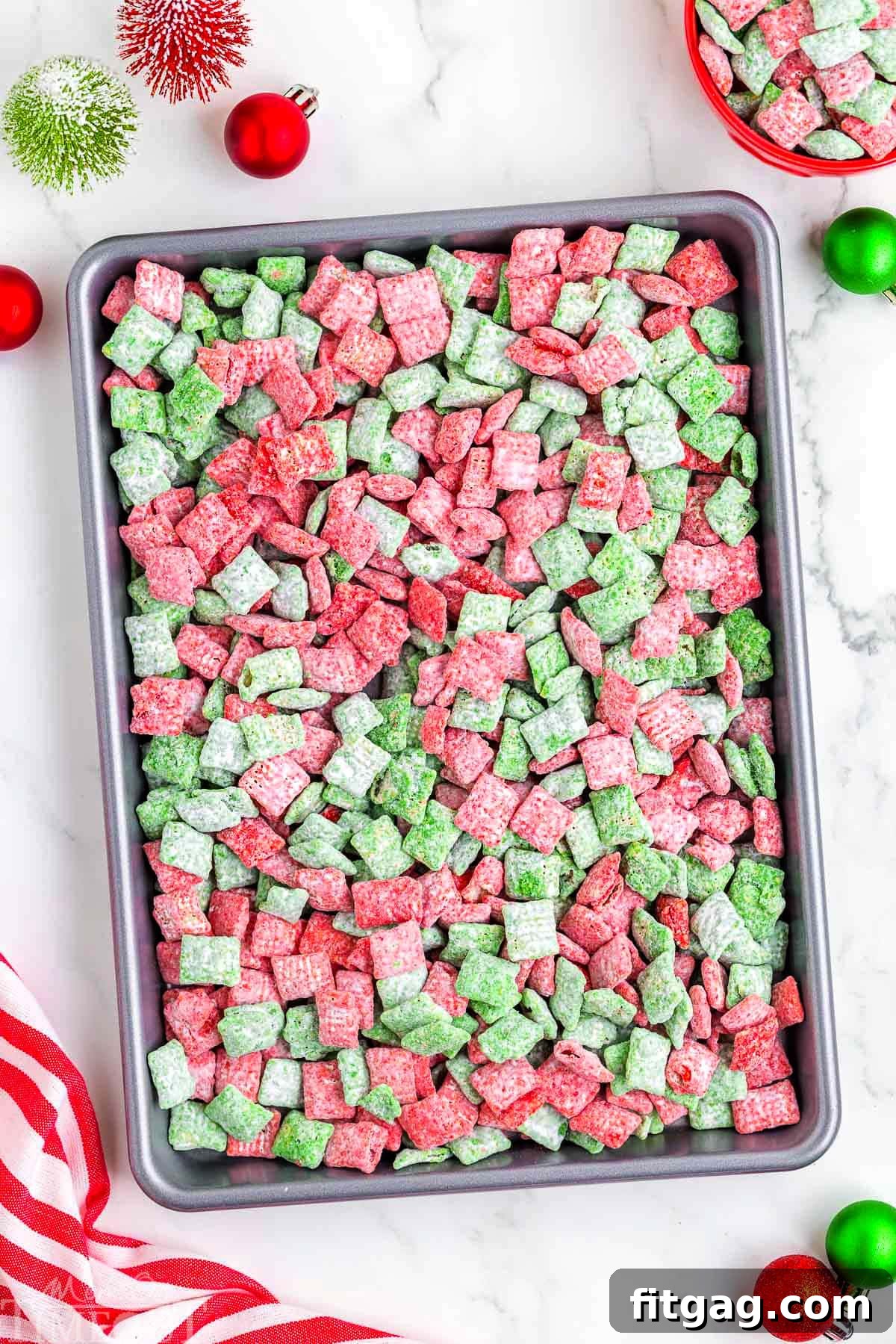 Holiday Crunch Mix 3 Overhead shot of freshly prepared Christmas Muddy Buddies spread evenly on a parchment-lined baking sheet, showing the distinct red and green coated cereal cooling and setting.