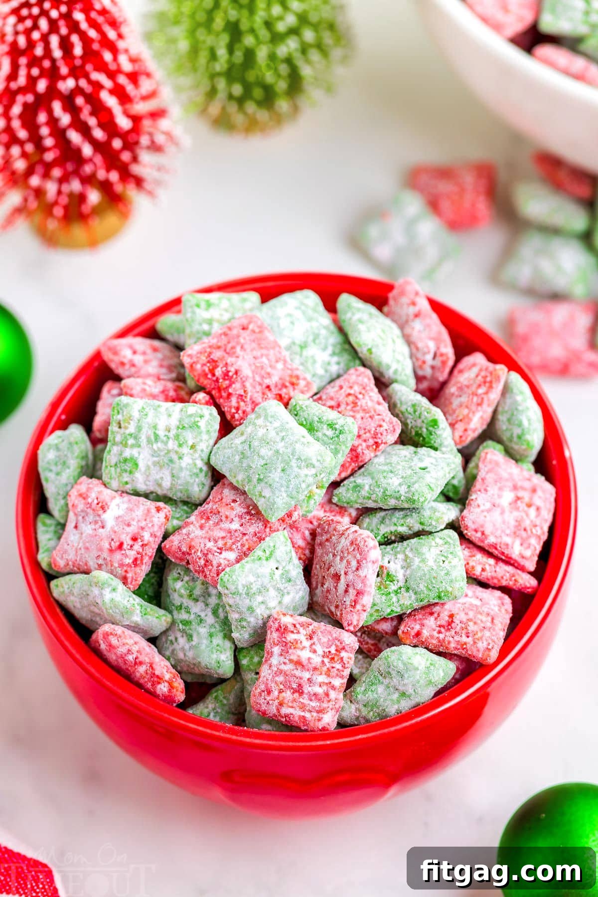 Holiday Crunch Mix 7 A vibrant red bowl filled to the brim with festive red and green Christmas Puppy Chow, placed on a crisp white surface with subtly blurred Christmas decorations in the background, evoking a warm holiday ambiance.