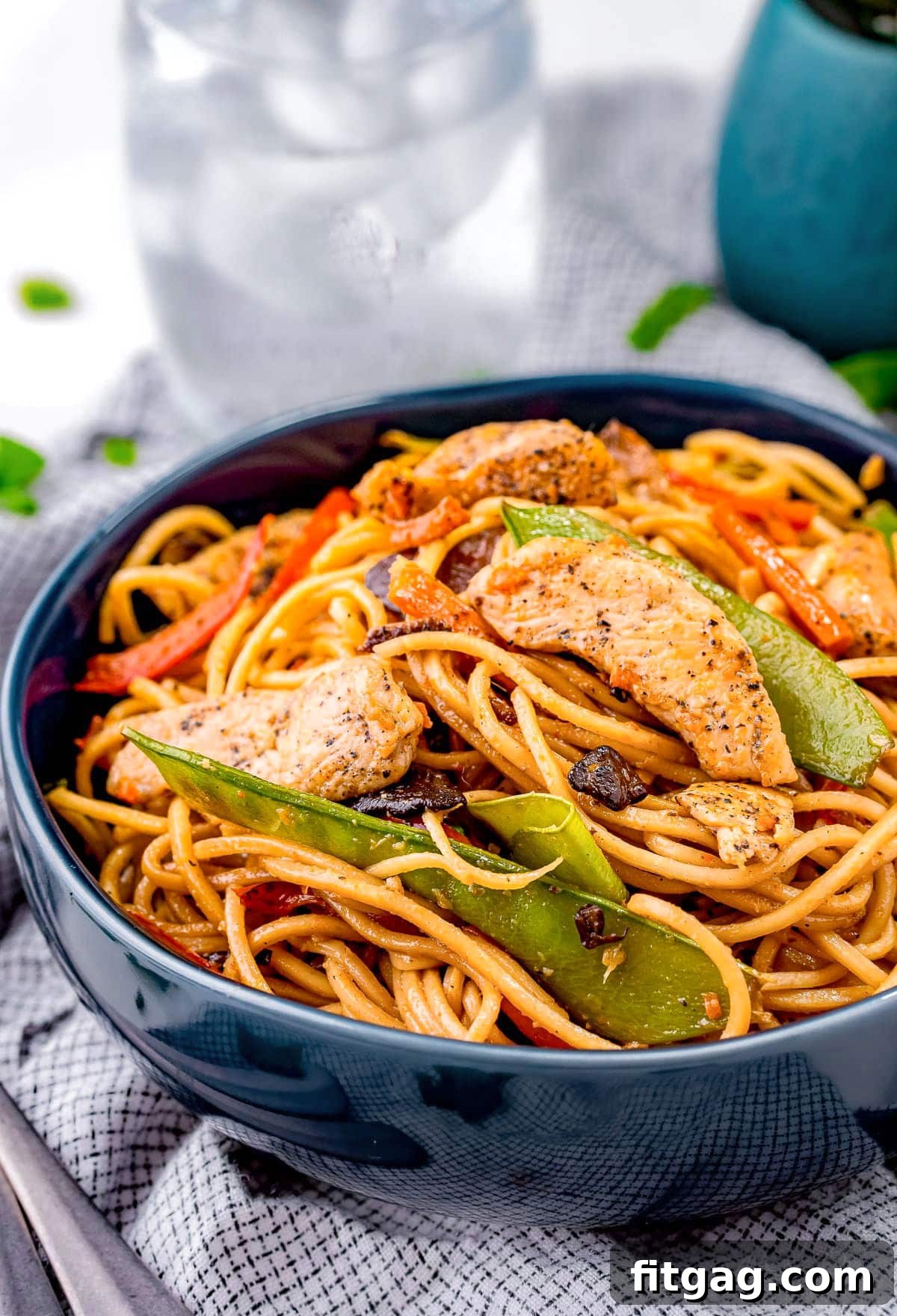 A vibrant blue bowl filled generously with homemade chicken lo mein, featuring tender chicken strips, green snow peas, and bright orange carrot shreds, garnished with fresh green onions.