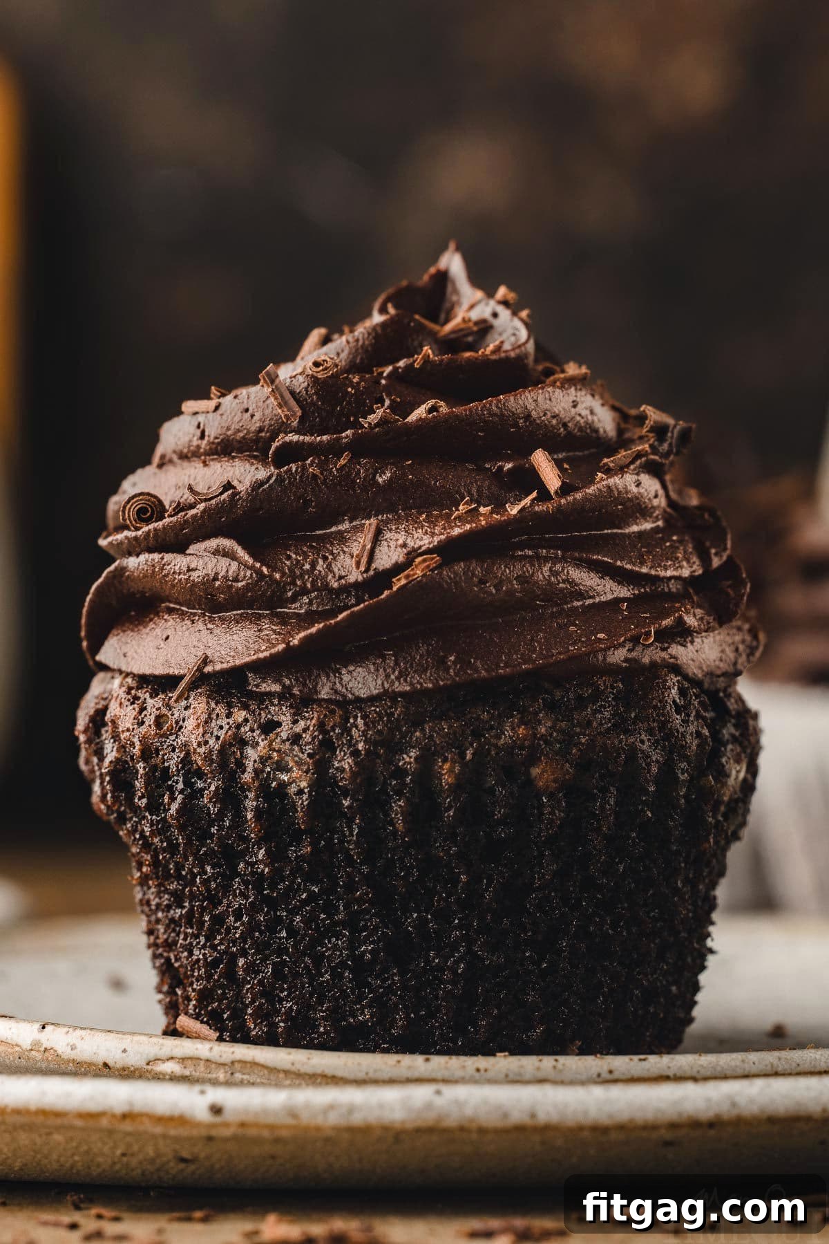 A perfectly moist chocolate cupcake, removed from its liner, sits on a white plate, beautifully topped with a silky chocolate buttercream frosting.