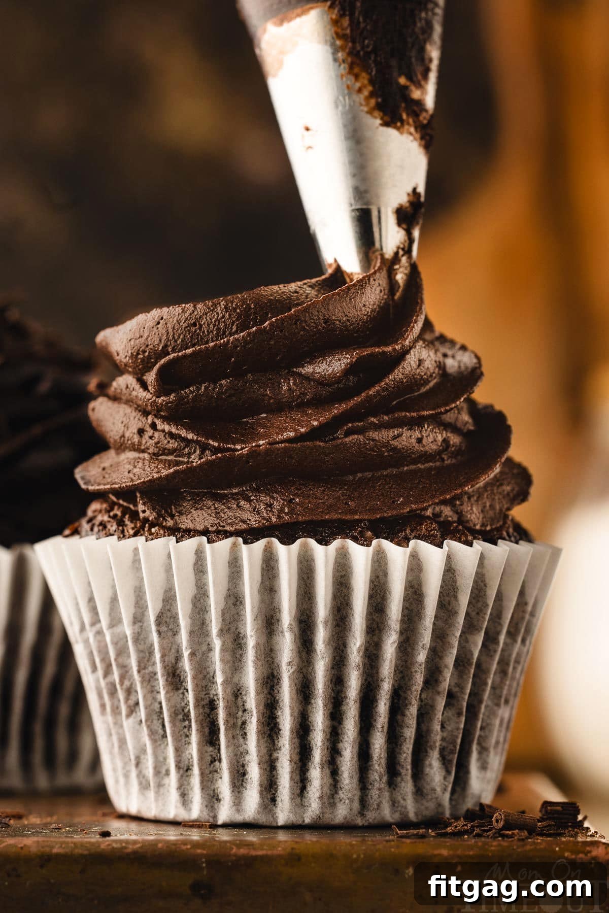 Chocolate buttercream being expertly piped onto a freshly baked chocolate cupcake, resting on a dark metal surface.