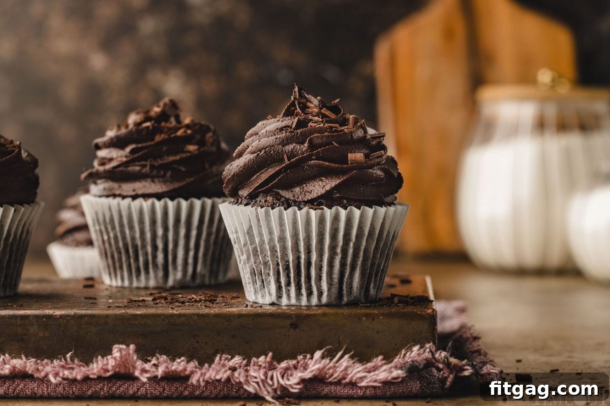 Two chocolate cupcakes in white liners topped with chocolate buttercream, with a cutting board visible in the softly blurred background.