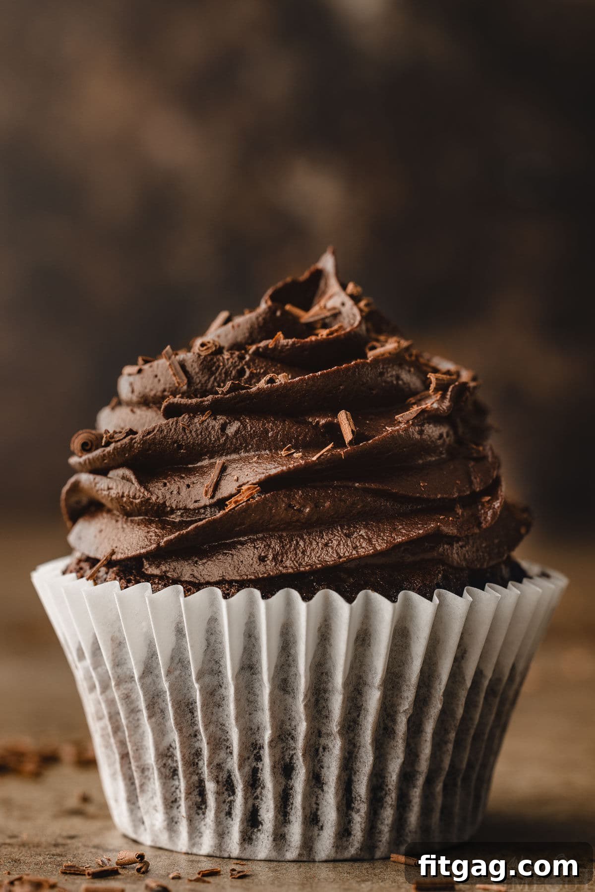 A perfect chocolate cupcake with deep chocolate buttercream still in its white wrapper, presented against a moody, dark background.