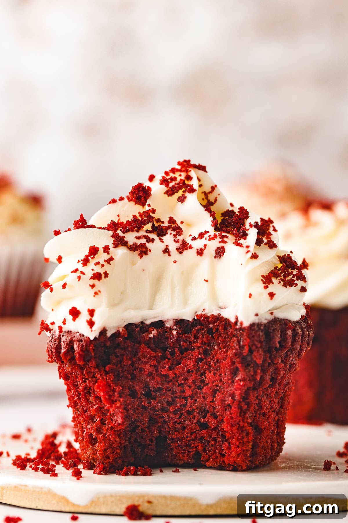 A single, perfectly frosted red velvet cupcake on a white ceramic plate, adorned with creamy cream cheese frosting and a sprinkle of red velvet crumbs. The vibrant red hue of the cupcake peeks through, promising a delightful treat.