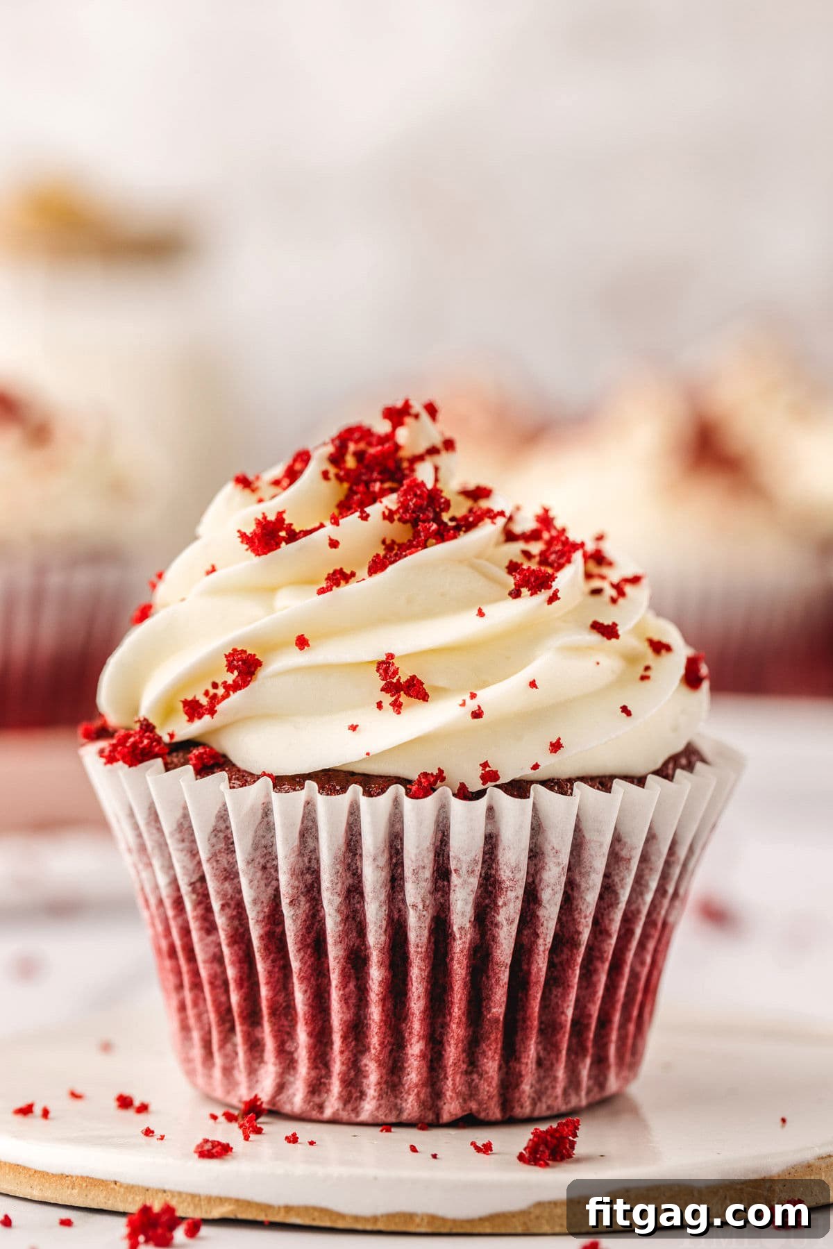 A beautifully presented red velvet cupcake in a white liner, resting on a white pottery plate. It's generously topped with velvety cream cheese frosting and a scattering of red velvet crumbs, with several other perfectly baked cupcakes blurred in the background, hinting at a celebratory spread.
