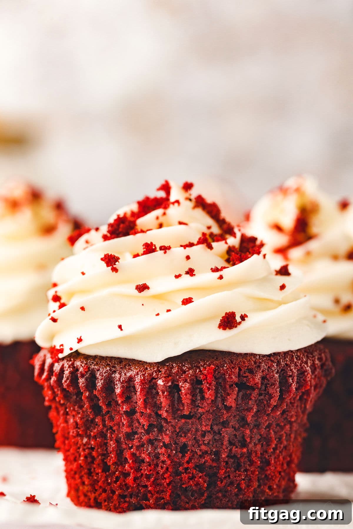 Another perspective of a single red velvet cupcake on a white pottery plate, featuring lush cream cheese frosting and a scattering of red velvet crumbs. More cupcakes are visible, creating a delightful sense of abundance.