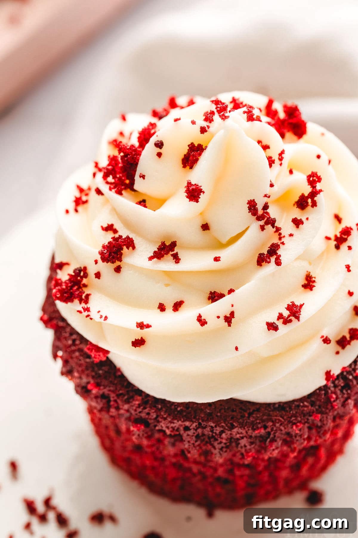 A top-down close-up shot of a red velvet cupcake, highlighting the rich, creamy texture of the cream cheese frosting. The frosting is beautifully swirled and sprinkled with red velvet crumbs, creating an inviting and irresistible appearance on a white plate.