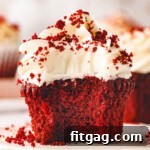 red velvet cupcake sitting on white pottery plate topped with velvety cream cheese frosting and sprinkle with red velvet crumbs.