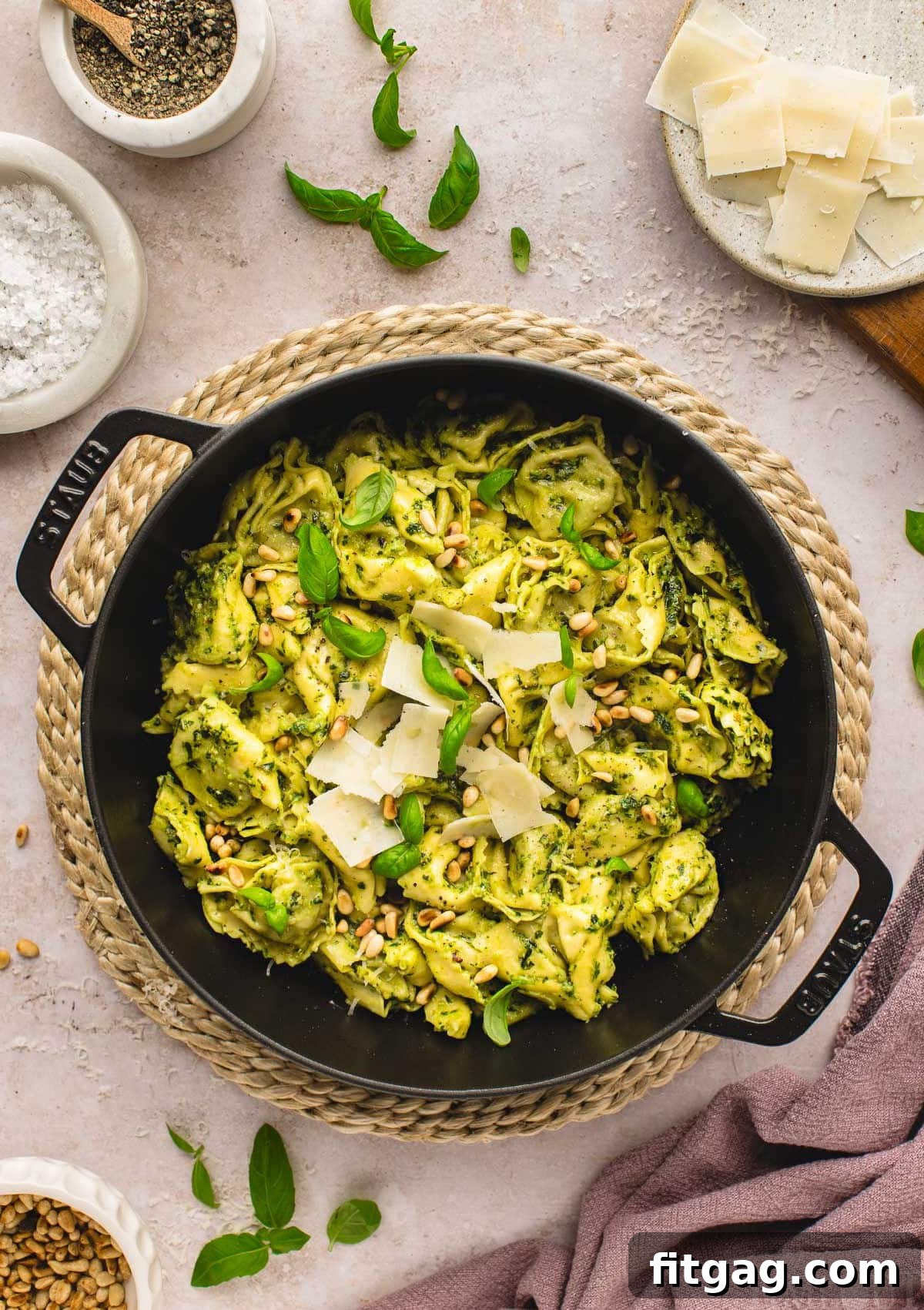 Creamy Pesto Tortellini Delight 7 top down shot of pesto tortellini recipe in large black cast iron skillet topped with shaved parmesan, toasted pine nuts and fresh basil leaves. small bowls of parmesan, salt and pepper can be seen around the skillet.