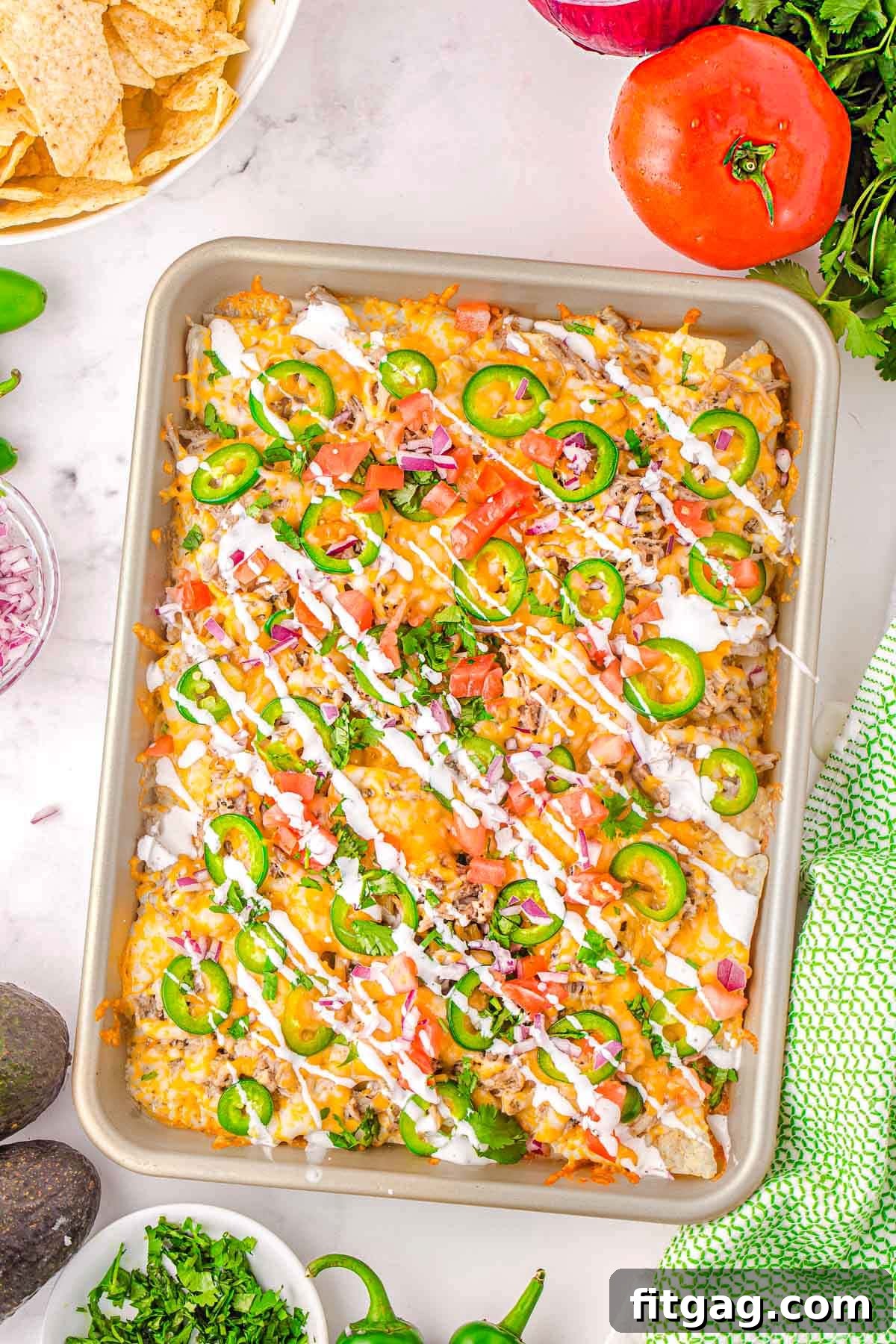 A top-down view of a large baking sheet generously filled with loaded pulled pork nachos, elegantly drizzled with sour cream and garnished with bright green cilantro, thinly sliced red onions, and fiery sliced jalapenos, all ready to be devoured.