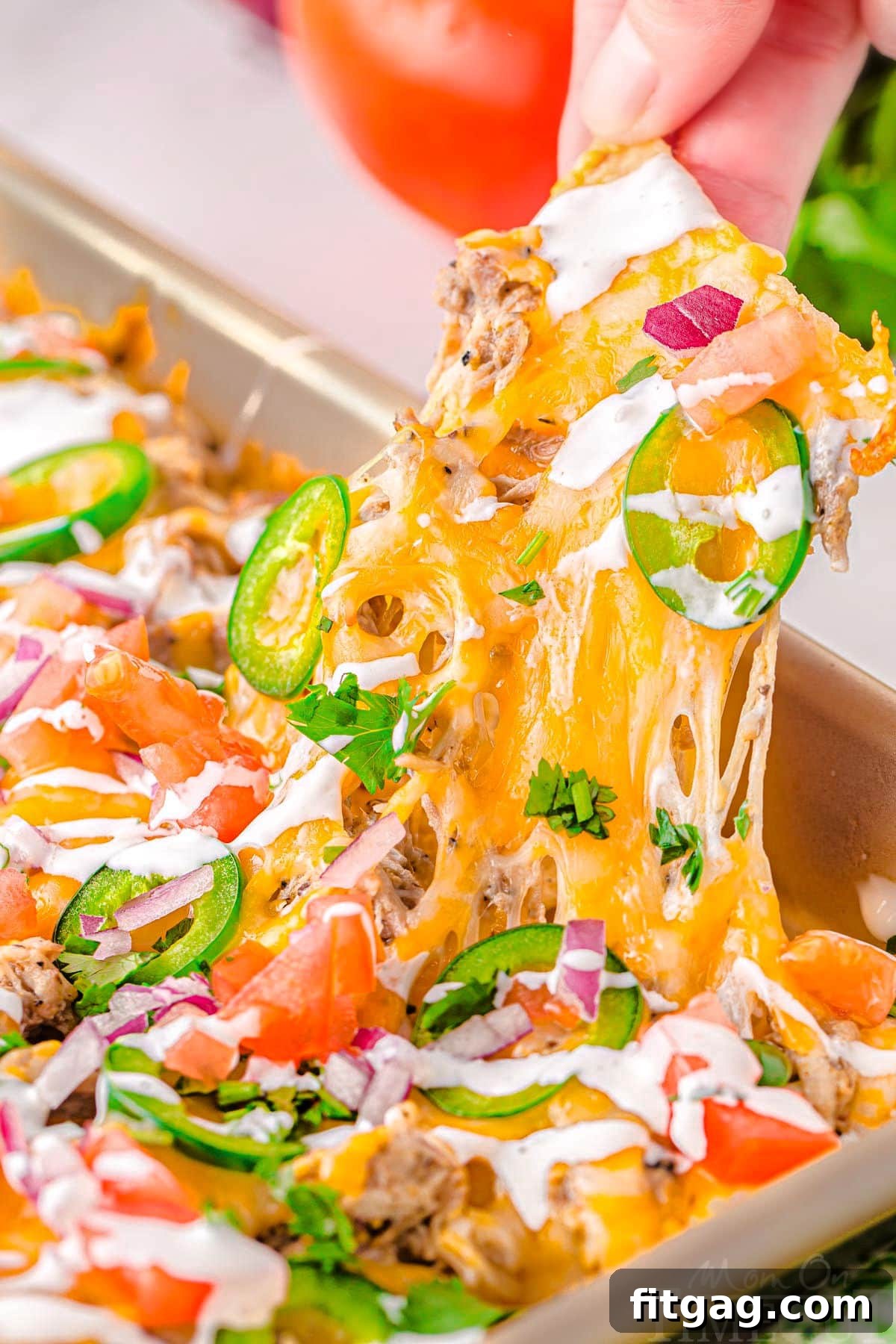 A hand gently lifts a portion of nachos from a large sheet tray filled with a vibrant spread of pulled pork nachos. The nachos are generously adorned with creamy crema, diced red tomatoes, and fresh green cilantro, showcasing their delicious layers and readiness for serving.