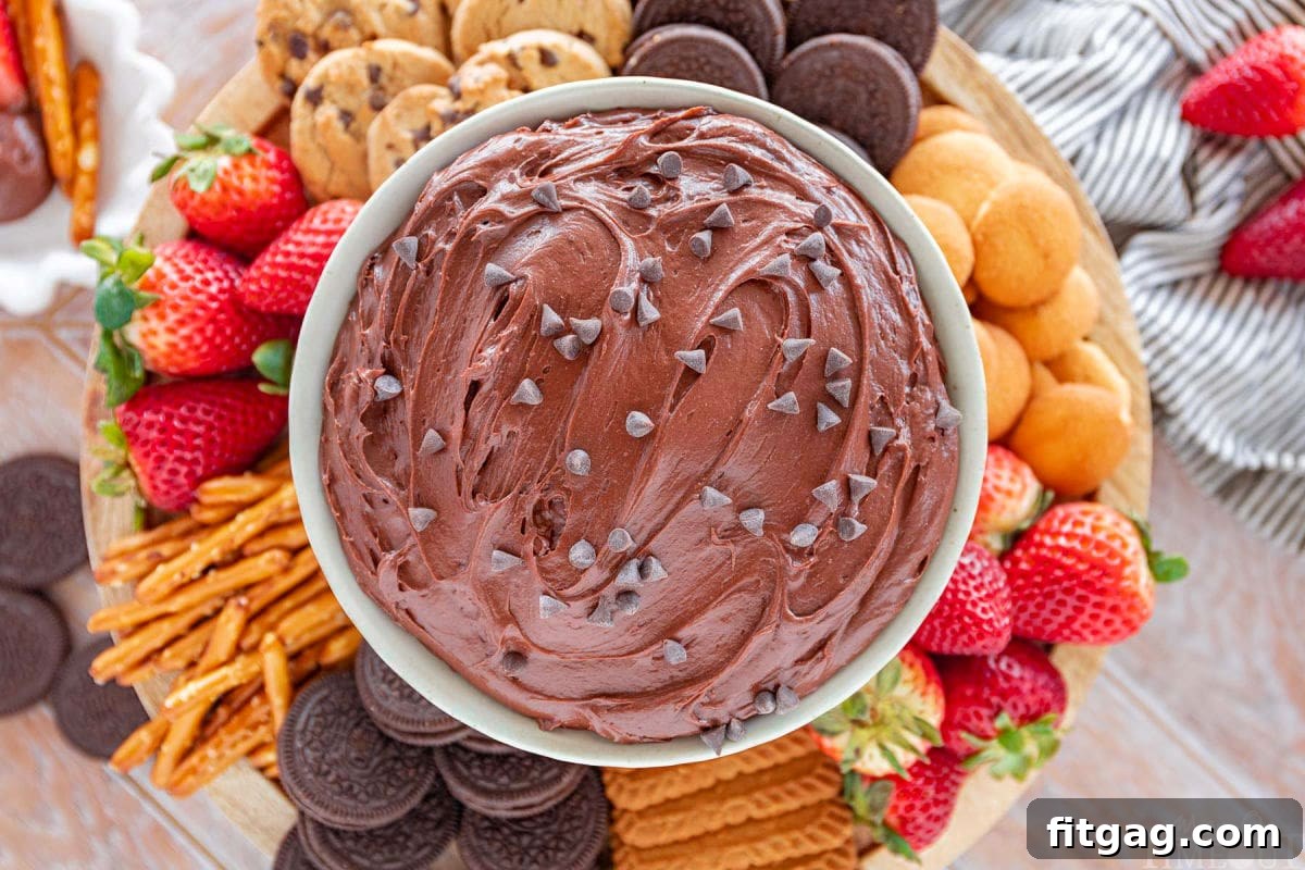 Fudgy Brownie Dip 5 Wide top-down shot of a large serving bowl filled with irresistible edible brownie batter dip, generously topped with mini chocolate chips, and elegantly surrounded by a colorful assortment of dippers including fresh strawberries, salty pretzels, an array of cookies, and other sweet treats.