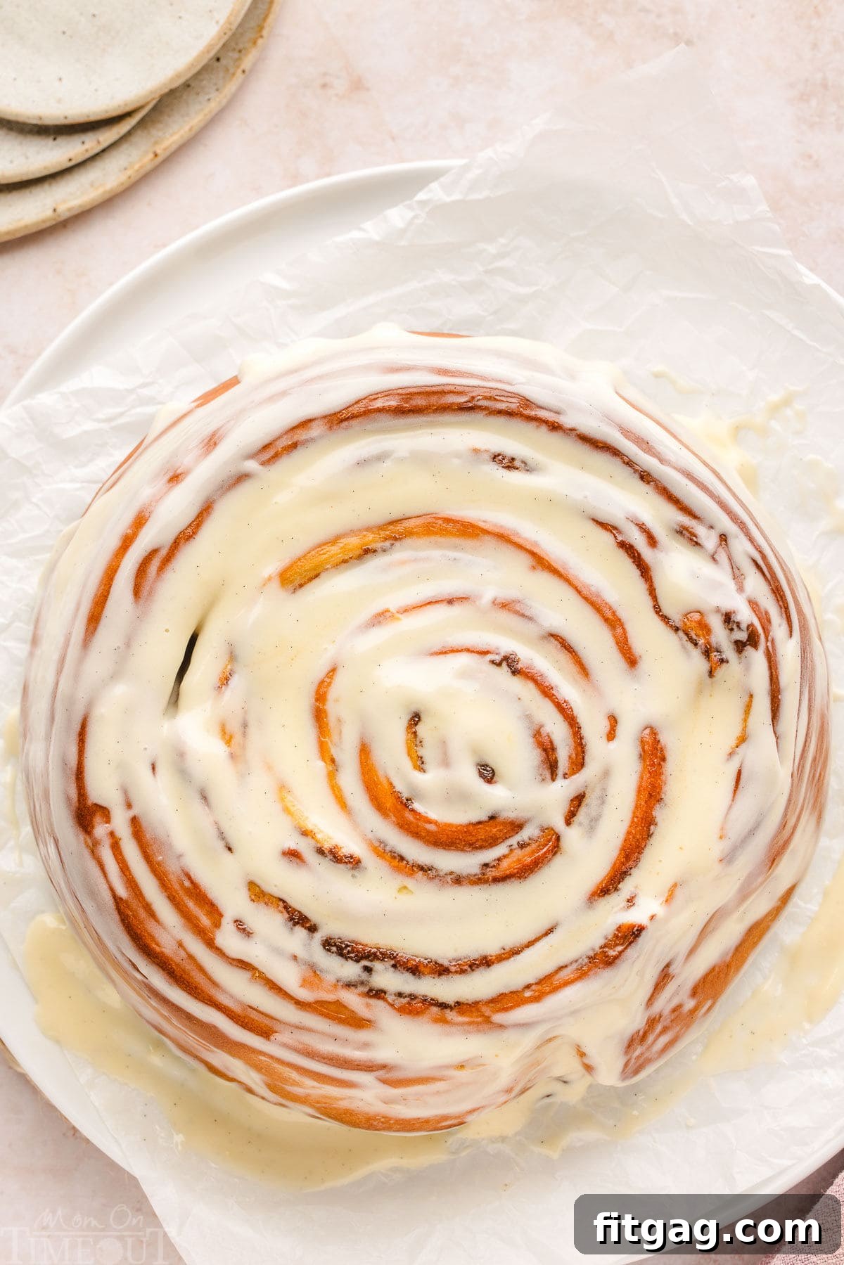 Colossal Cinnamon Swirl Cake 4 giant cinnamon roll with a cream cheese glaze sitting on white parchment with small plates to the side.
