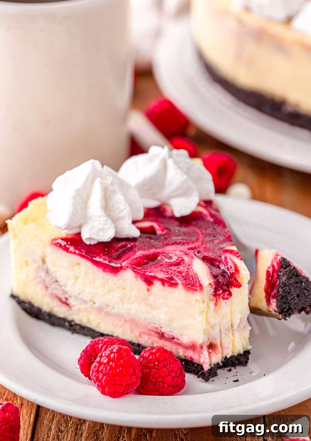 Velvet White Chocolate Raspberry Bliss 2 A perfectly sliced piece of white chocolate raspberry cheesecake on a round white plate, elegantly topped with a dollop of fresh whipped cream and three ripe raspberries. The layers of the cheesecake are visible, showcasing the crunchy Oreo crust, creamy white chocolate filling, and vibrant raspberry swirl.