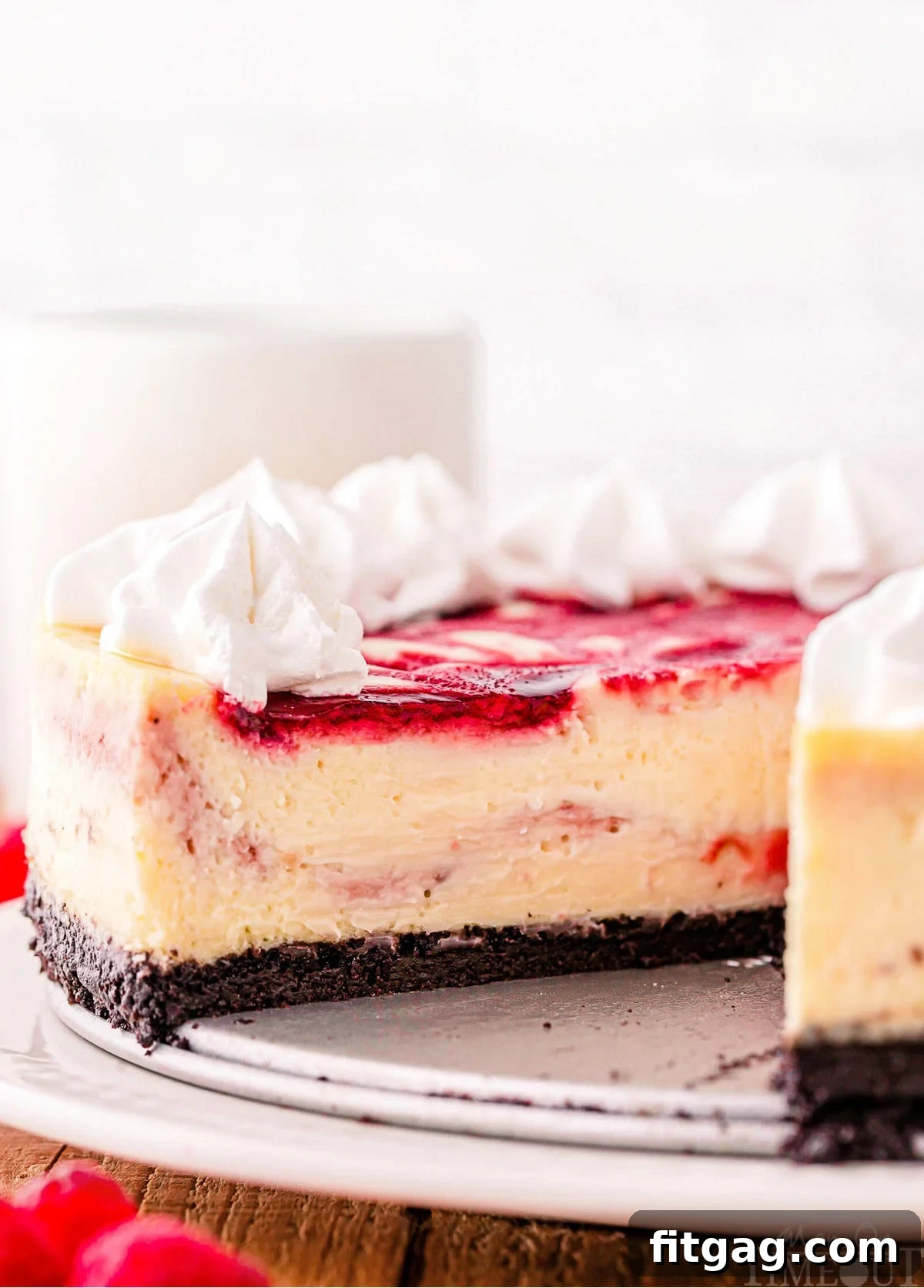 Velvet White Chocolate Raspberry Bliss 3 A beautiful profile shot of the White Chocolate Raspberry Cheesecake, highlighting its distinct layers: a dark, crunchy Oreo crust, a thick, creamy white chocolate filling, and a visible raspberry swirl, all topped with a generous amount of fluffy whipped cream and fresh raspberries.
