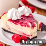 slice of white chocolate raspberry cheesecake on a round white plate topped with whipped cream. three raspberries are sitting next to the cheesecake on the plate.