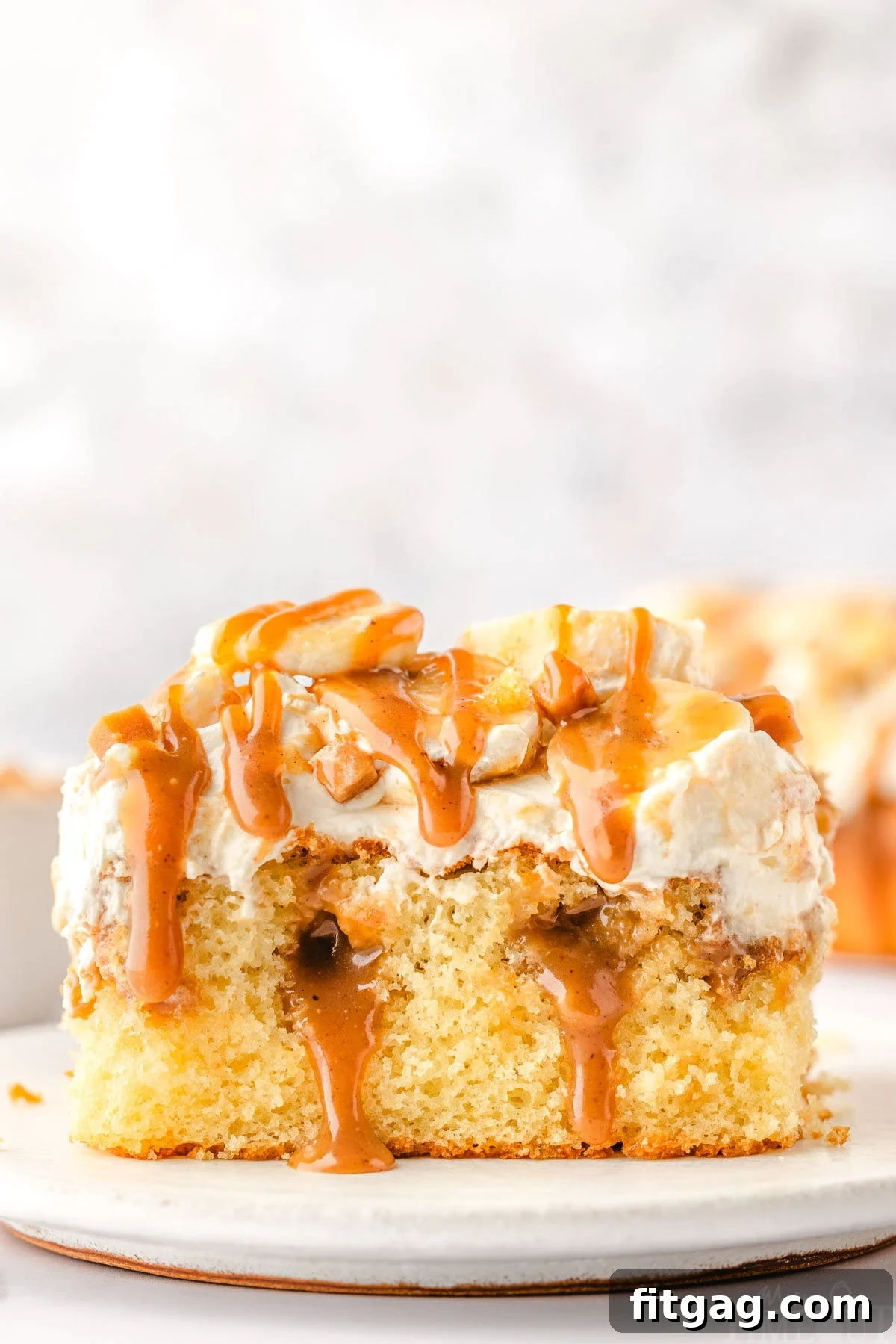 A perfectly sliced piece of Banoffee Poke Cake on a white plate, showcasing layers of yellow cake, golden dulce de leche, fluffy whipped cream, and fresh banana slices, garnished with a drizzle of caramel.