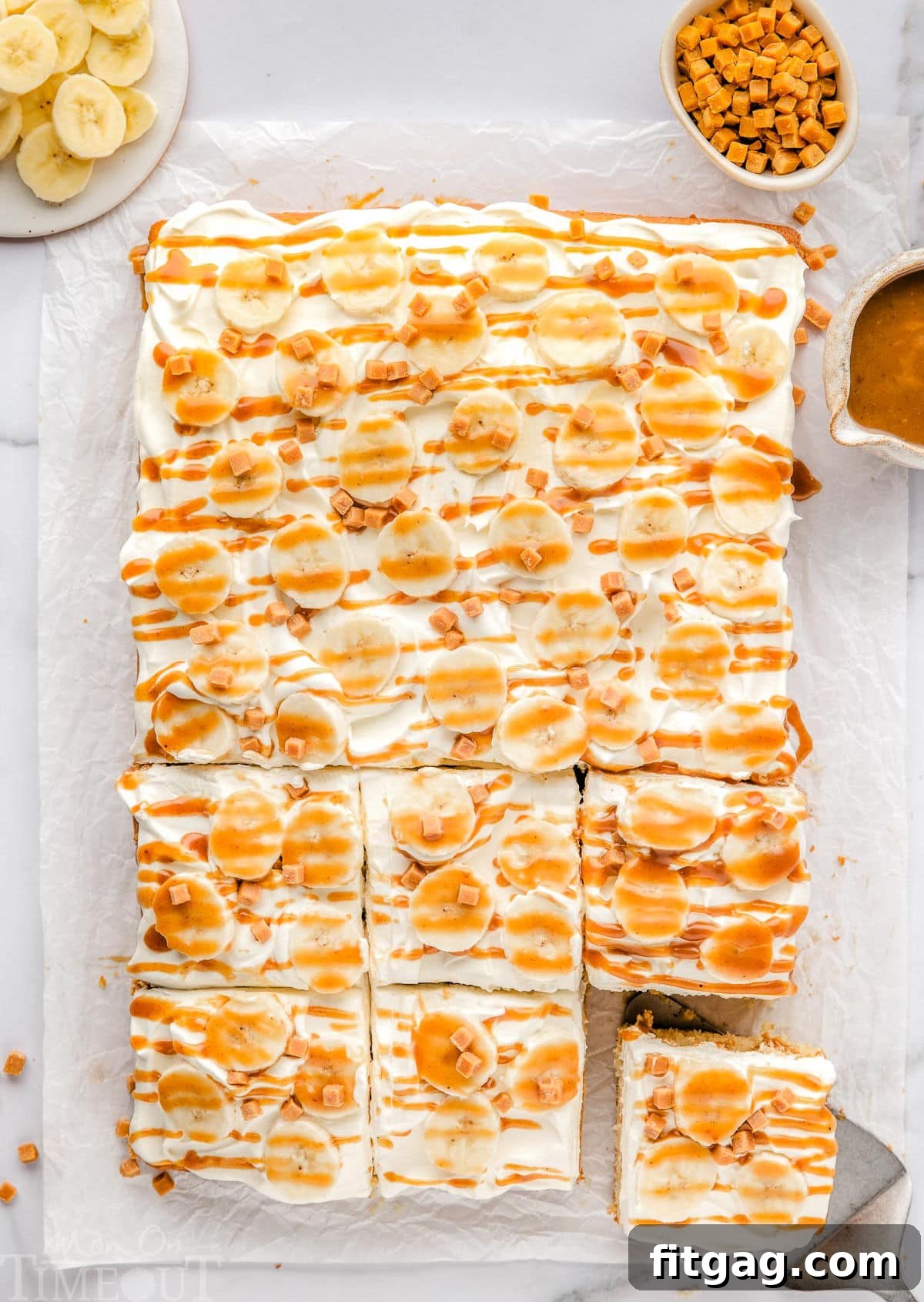 An overhead view of a freshly baked Banoffee Poke Cake, with several pieces already cut, resting on white parchment paper. A bowl of sliced bananas and toffee bits is positioned next to the cake, ready for serving or garnishing.