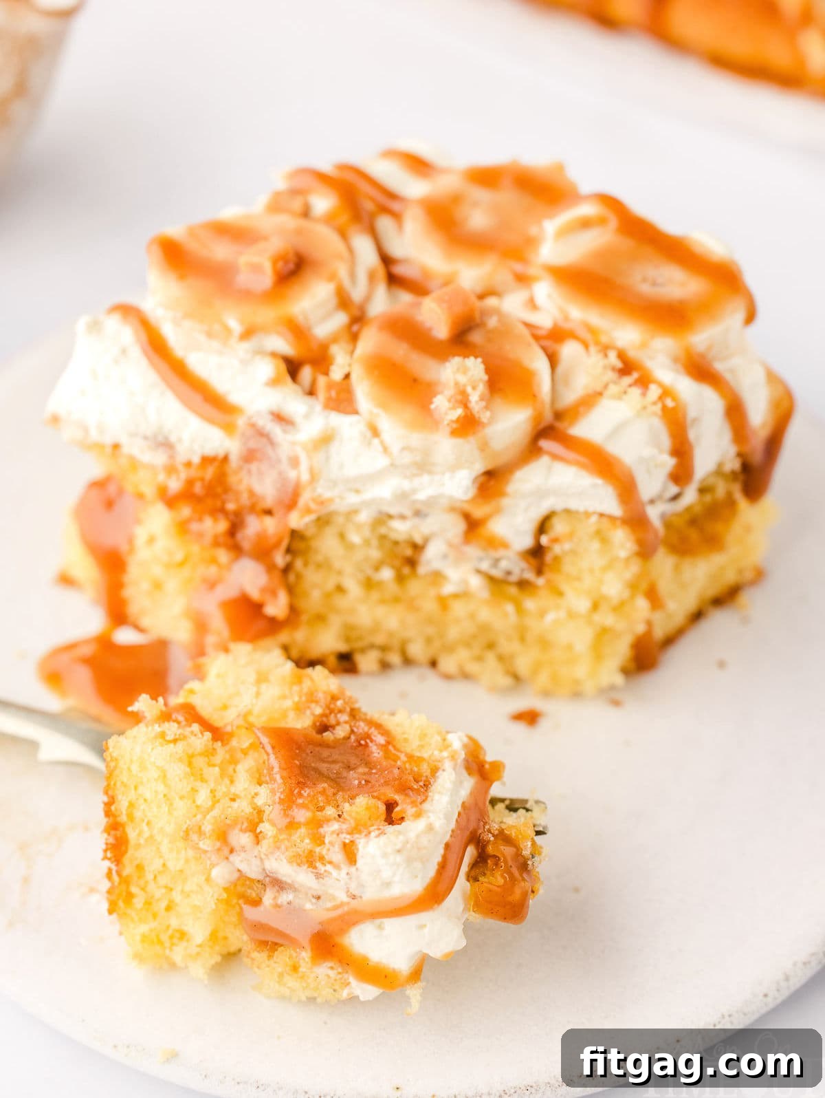 A fork taking a bite of Banoffee Poke Cake, showing the moist cake, dulce de leche, whipped cream, and bananas on a small, round white plate beside the rest of the slice.