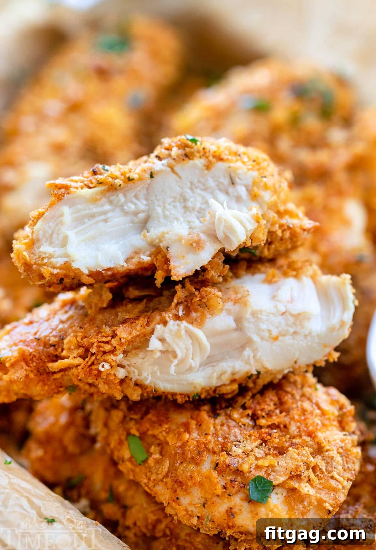 A pile of golden-brown Cornflake Chicken tenders with a side of creamy ranch dipping sauce. One tender is cut in half, revealing the moist, juicy chicken interior and the perfectly crisp, flavorful cornflake coating.