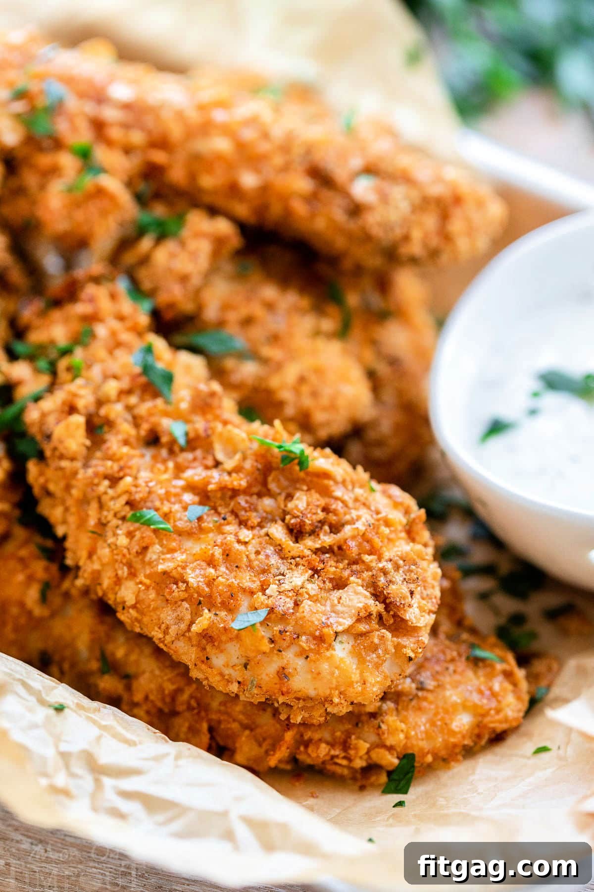 A small baking sheet lined with brown parchment paper, overflowing with a delicious pile of crispy Cornflake Chicken strips. A bowl of creamy ranch dressing is ready for dipping next to the chicken.