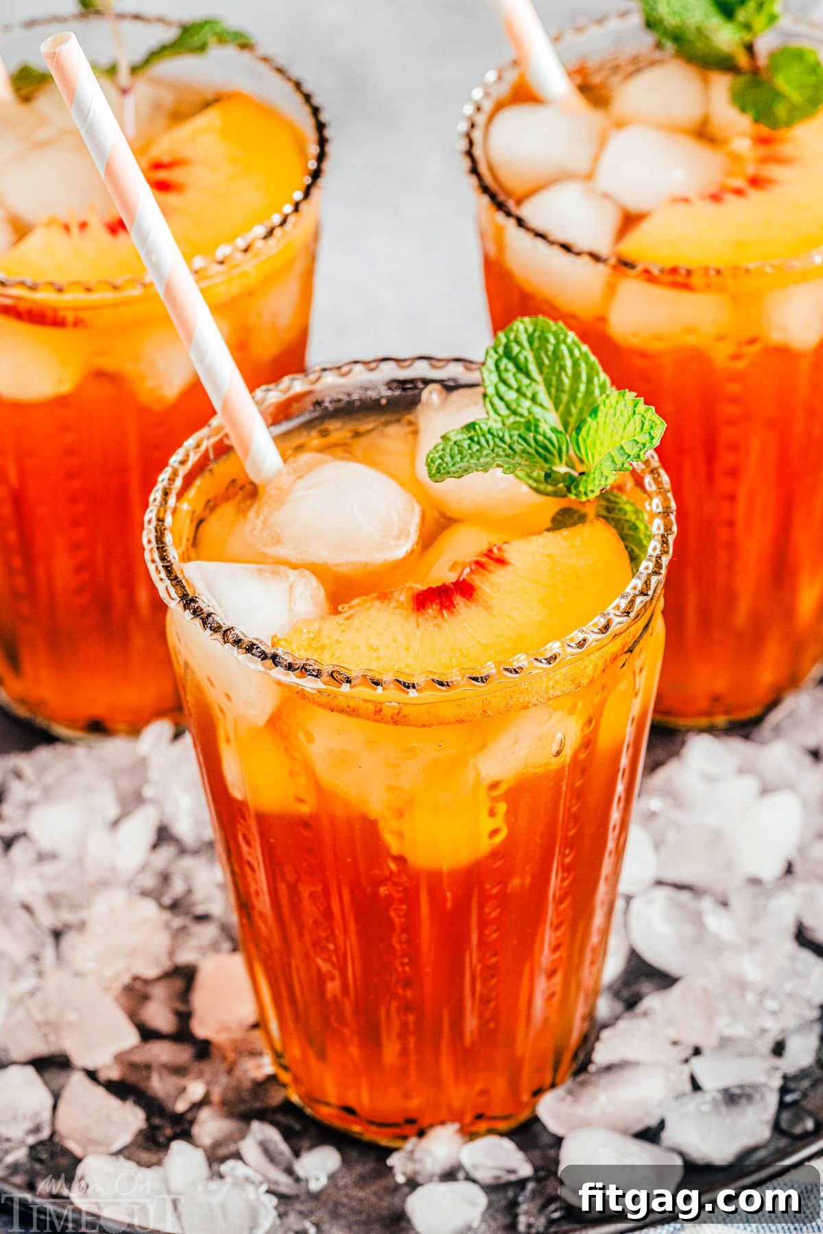 Sun-Kissed Peach Brew 2 three tall glasses filled with homemade peach iced tea and topped with fresh peach slices and a sprig of mint. glasses are sitting on a tray covered with ice.