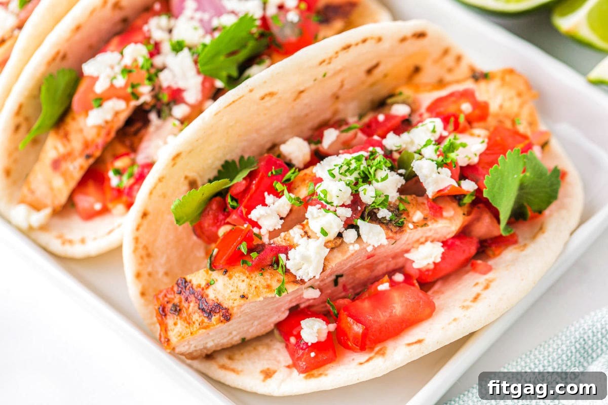 three chicken tacos on a white rectangular plate topped with pico de gallo, cilantro and queso fresco. A half a lime can be seen in the background.