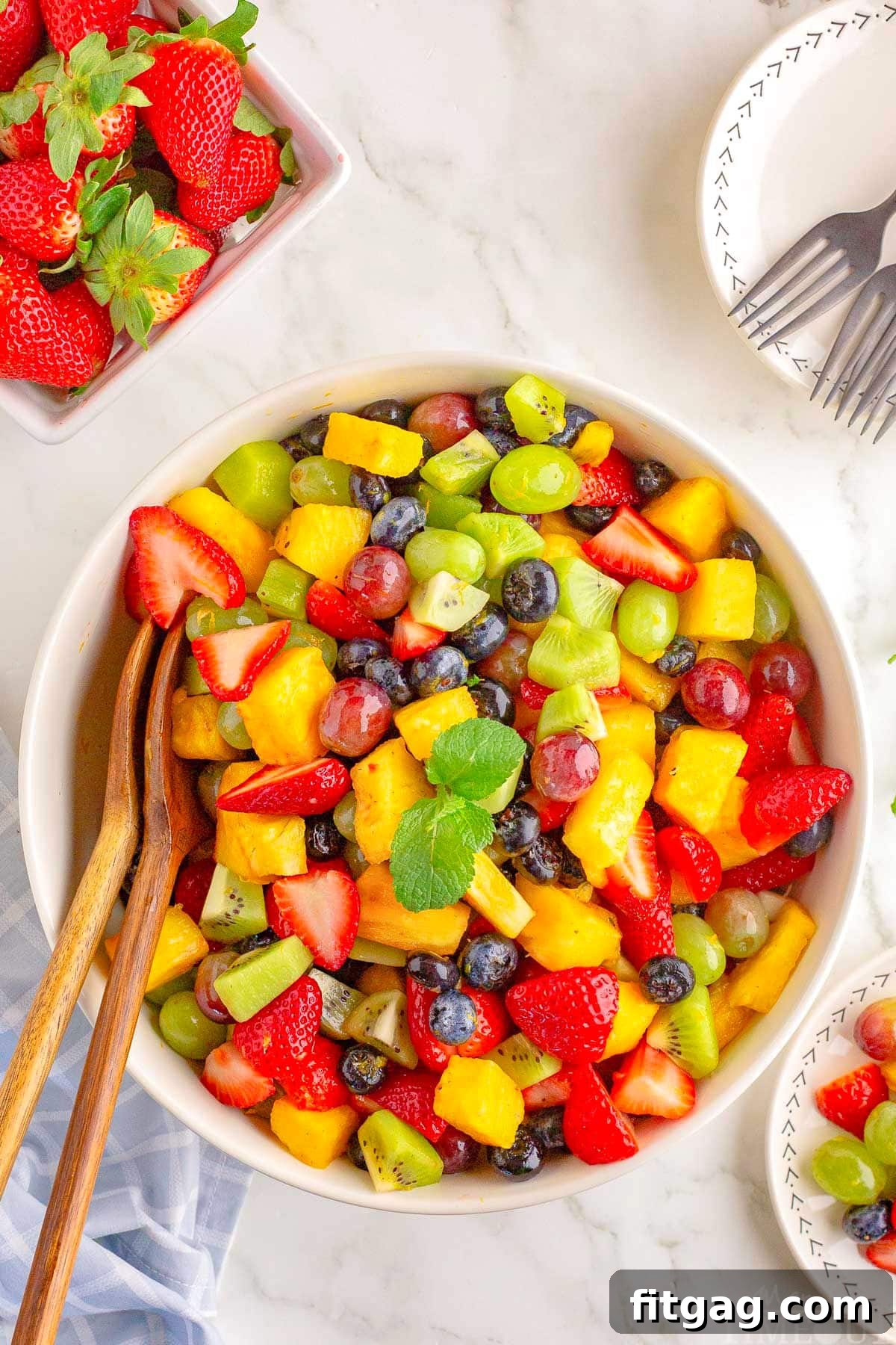 Sunshine Fruit Medley 2 A vibrant bowl of fresh fruit salad, featuring pineapple, strawberries, blueberries, kiwi, and grapes, with wooden salad tongs ready for serving.