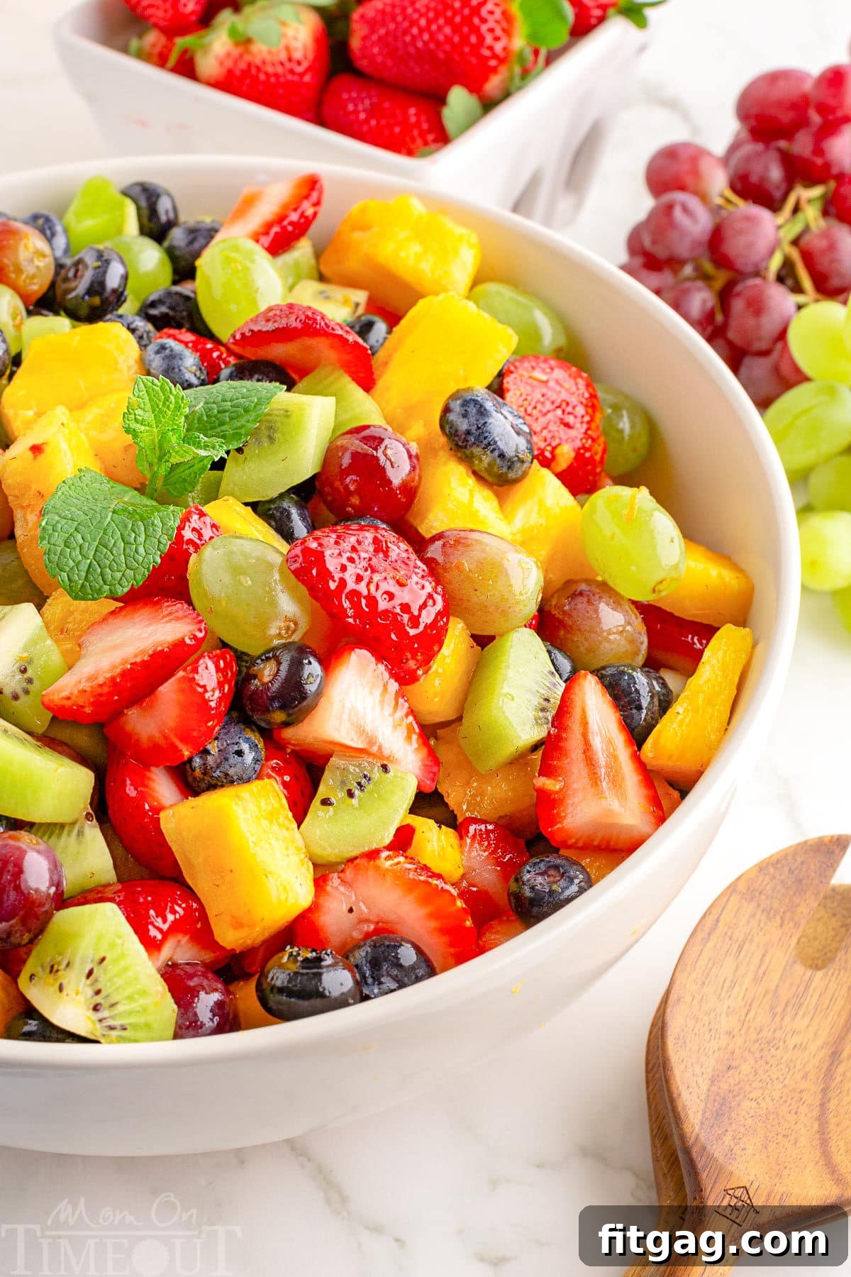 A white bowl of fruit salad with fruit in the background.