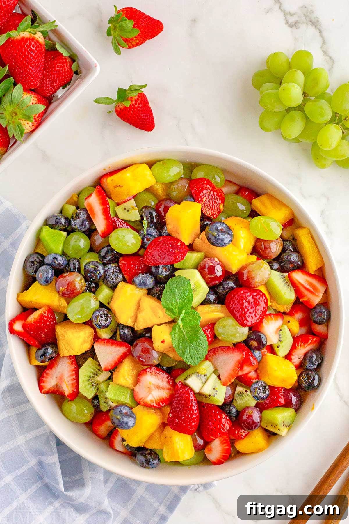 top down shot of fresh fruit salad in large white serving bowl topped with a sprig of mint. Green grapes and strawberries are scattered around the outside of thr bowl.